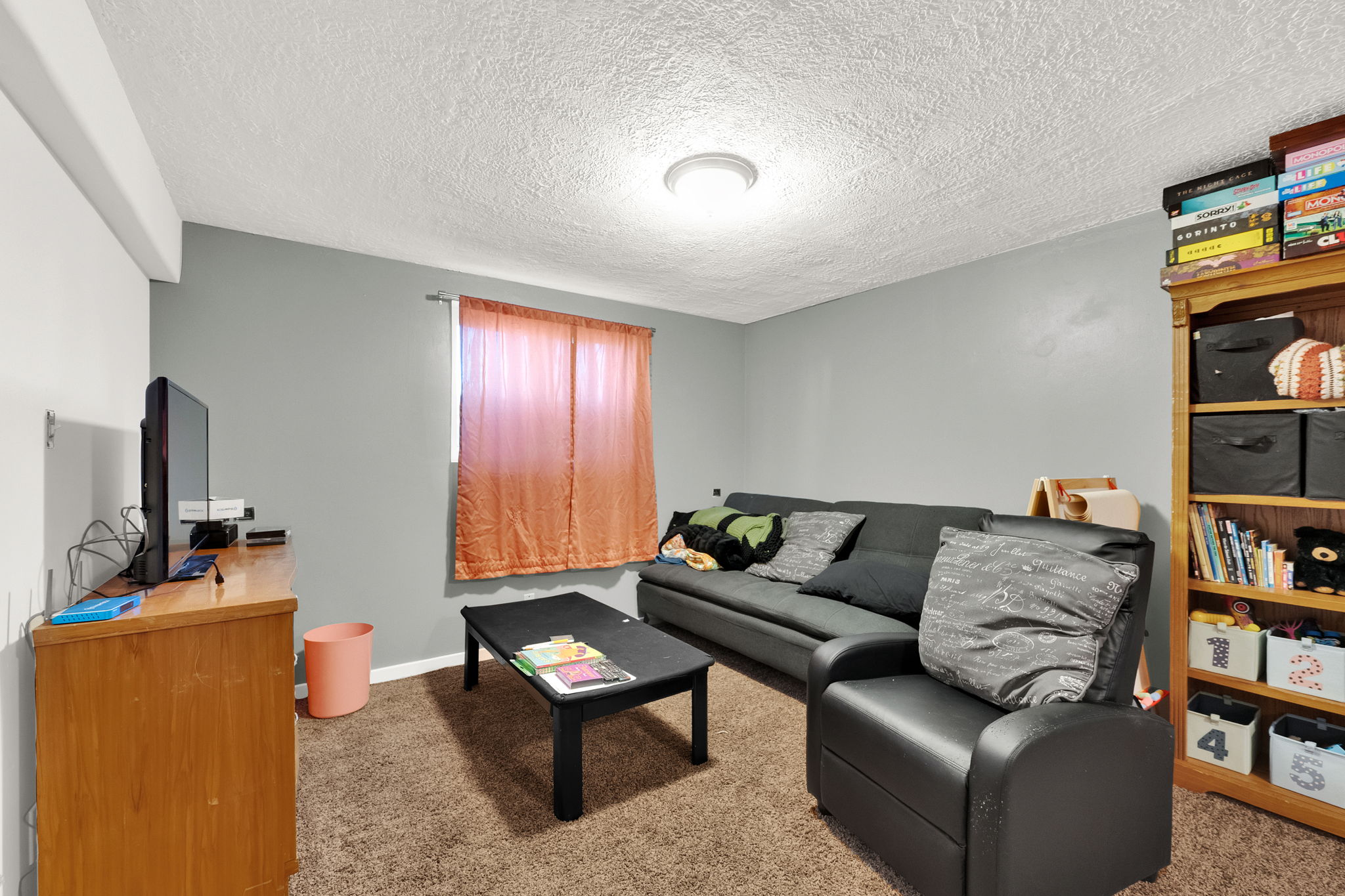 Carpeted room with a ceiling-mounted light fixture, neutral gray walls, and a window with light-filtering curtains