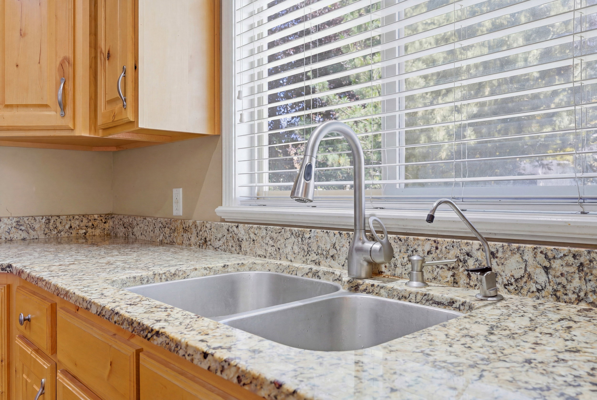 Kitchen sink area featuring a double basin stainless steel sink with a gooseneck faucet, light granite countertops, natural wood cabinetry, and a window with horizontal blinds