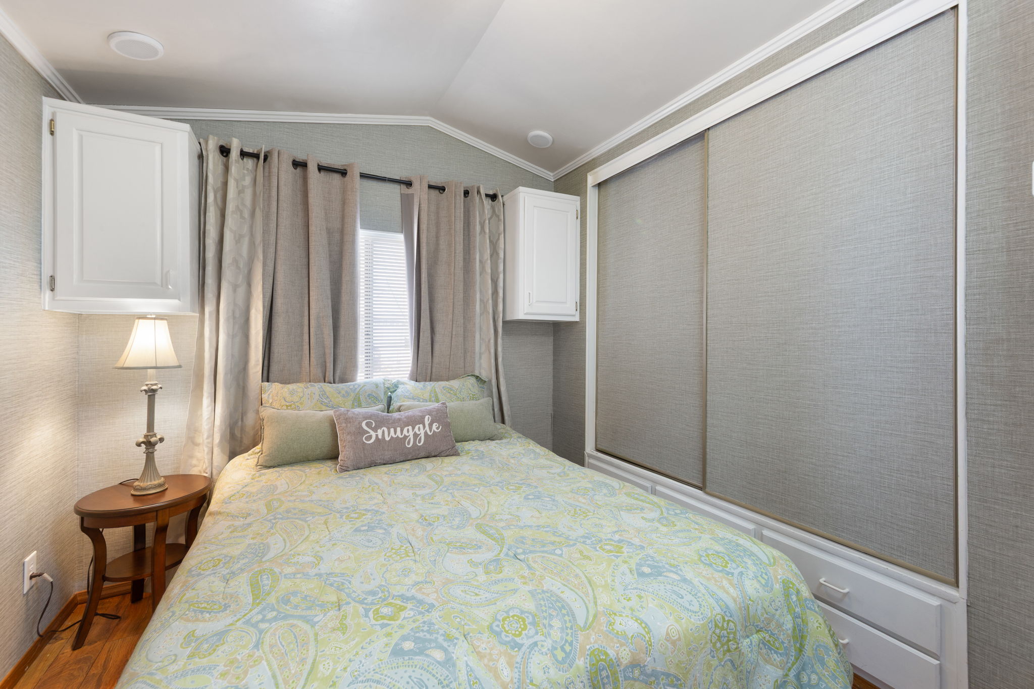 Bedroom featuring wood-finish flooring, two white wall-mounted cabinetry units, a window with horizontal blinds and drapery, and a built-in closet with sliding doors and integrated drawers