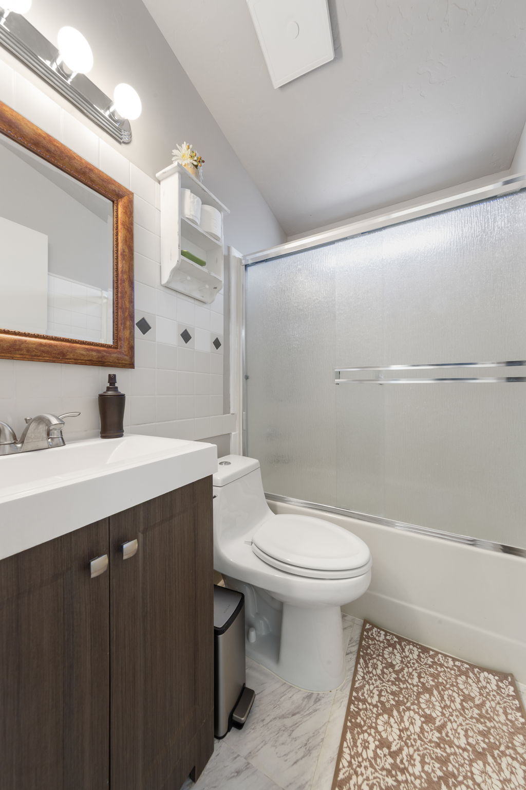 Bathroom featuring a dark wood-finish vanity with a white countertop and rectangular sink, a white toilet, and a shower-tub combination with a frosted sliding glass enclosure