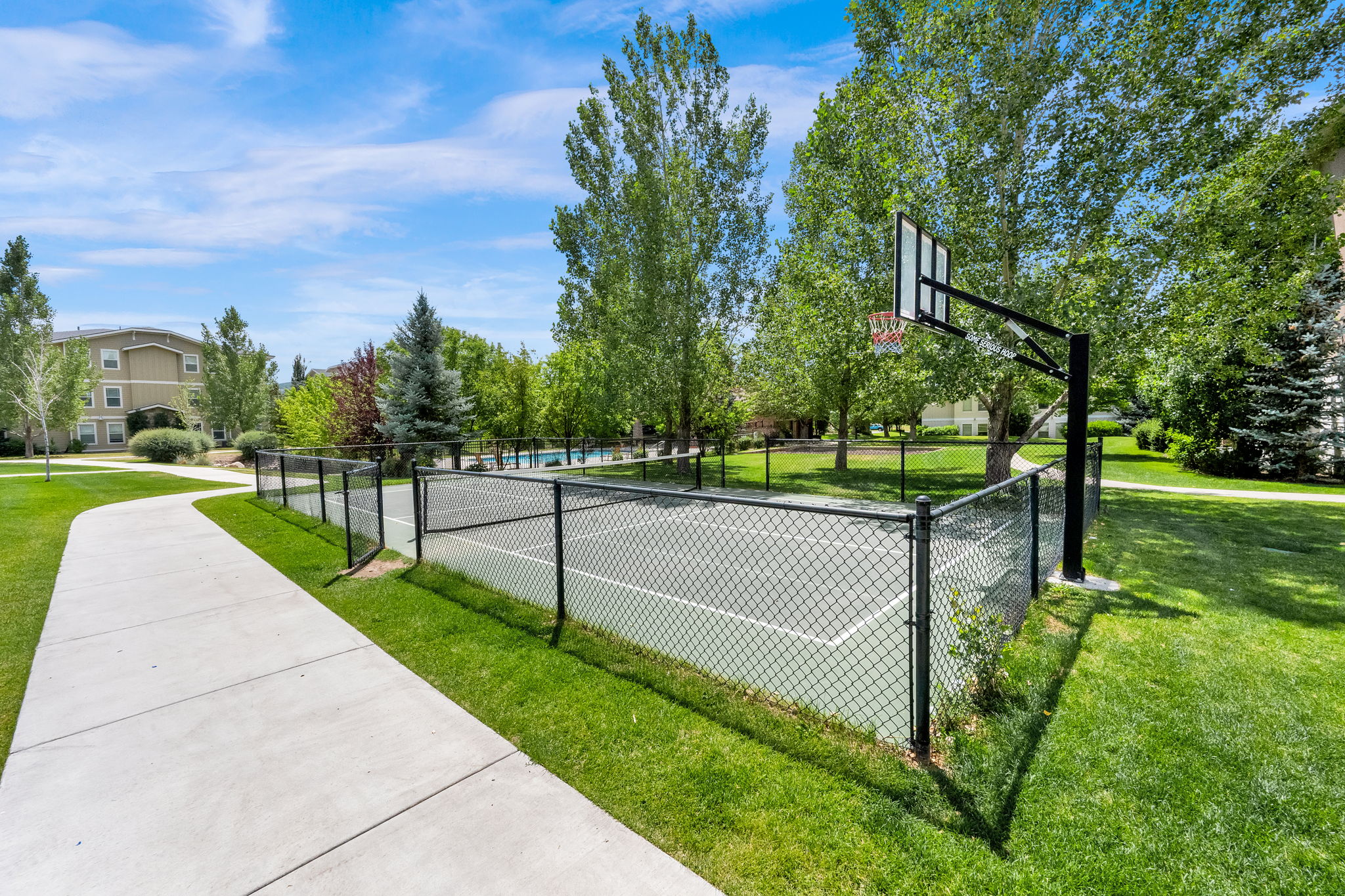 Outdoor basketball court with chain-link fencing, surrounded by mature trees and manicured lawns
