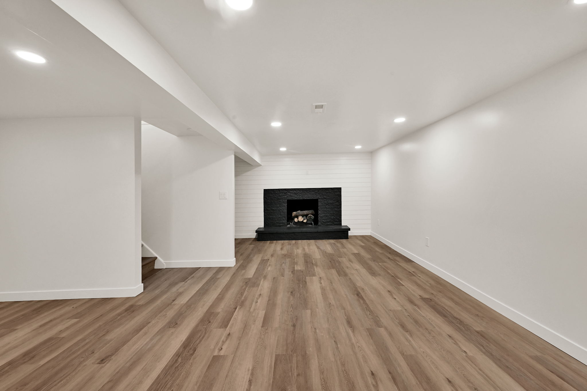 Spacious room featuring wood-finish flooring, a black stone-clad fireplace with hearth, white shiplap accent wall, recessed lighting, and white baseboards