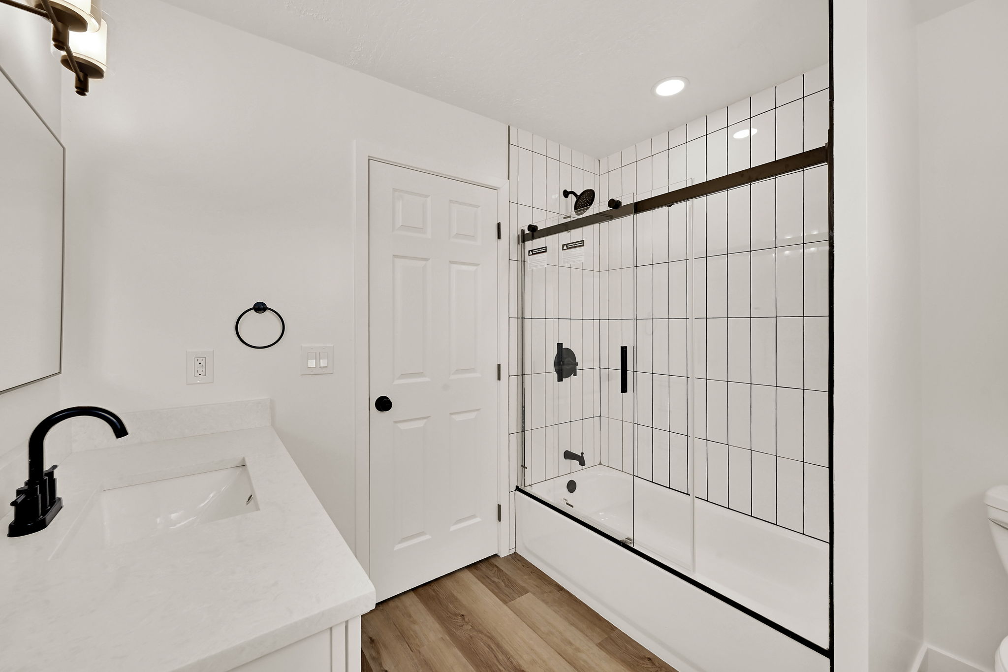 Bathroom featuring a white subway tile shower surround with black hardware, a shower/tub combination with a sliding glass door, a vanity with an integrated sink and light-toned countertop, black faucet, and wood-finish flooring