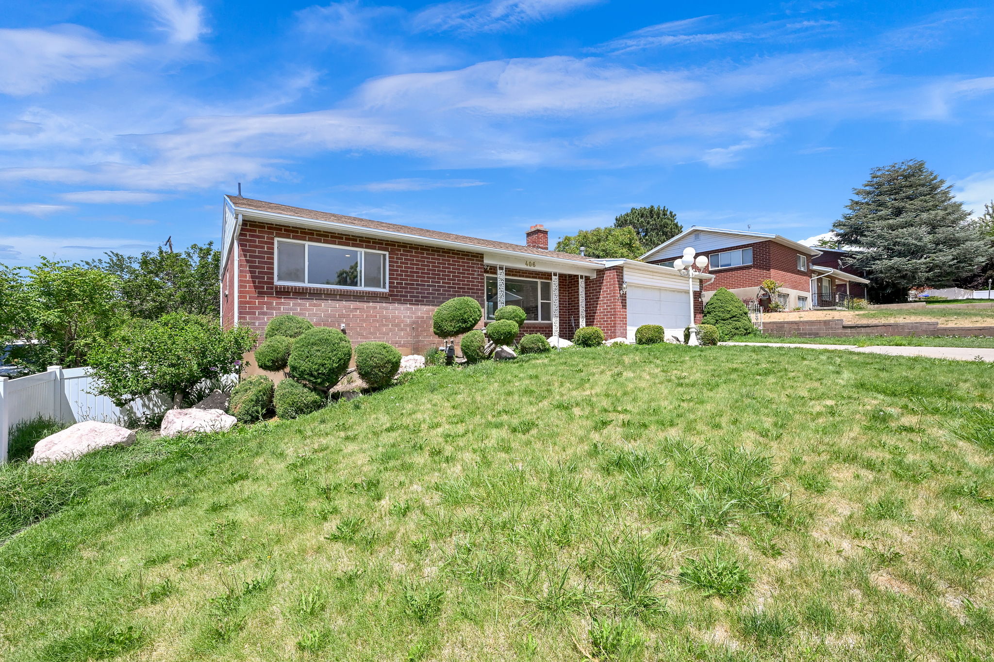 Brick exterior home featuring a sloped front lawn with mature shrubbery and a white privacy fence