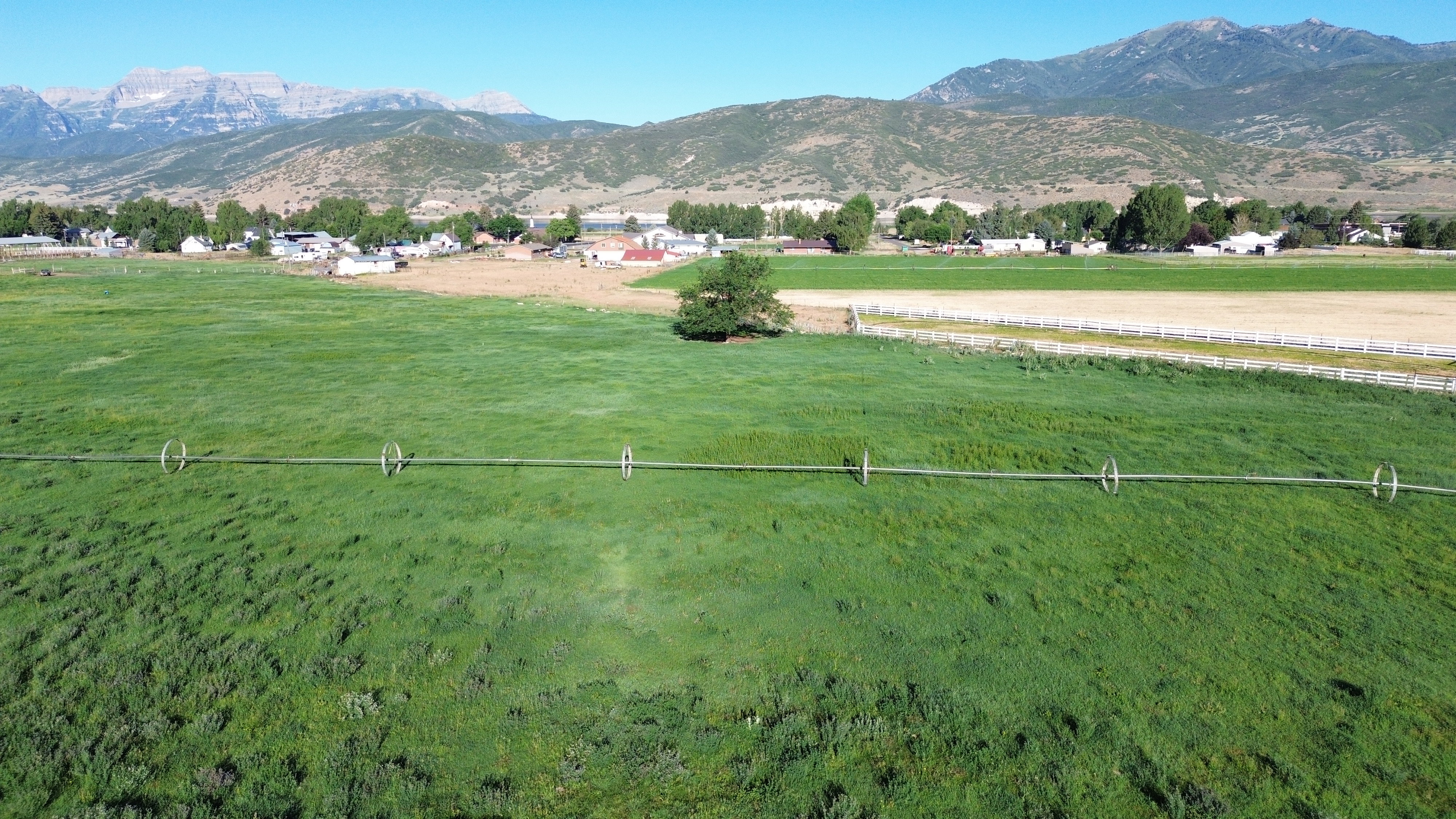 Expansive green field featuring an irrigation system, bordered by a white fence and a distant community, with mountains forming a backdrop