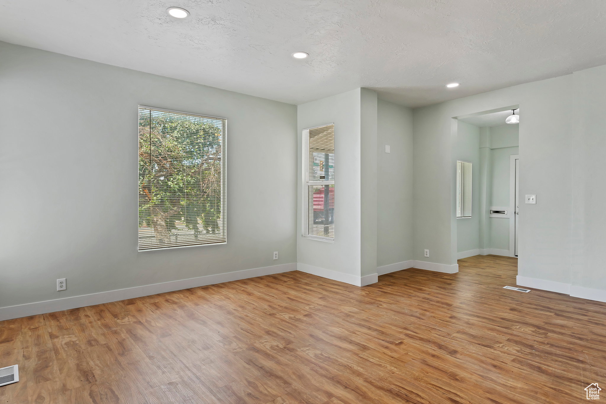 Spacious interior room featuring wood-finish flooring, two large windows with natural light, recessed ceiling lighting, and a neutral wall color palette