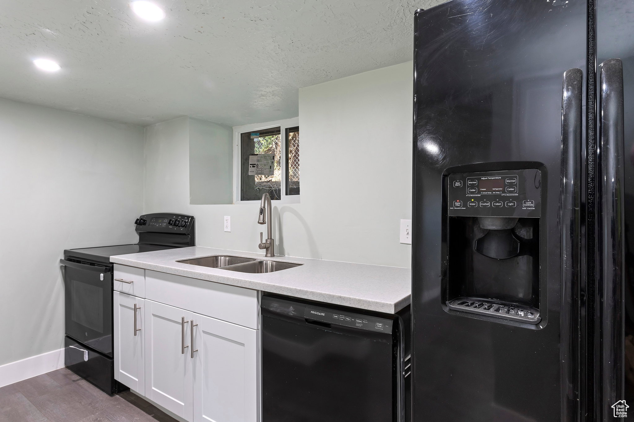 Modern kitchen featuring a black refrigerator with ice and water dispenser, stainless steel sink with gooseneck faucet, white shaker-style cabinetry, and light-toned countertops