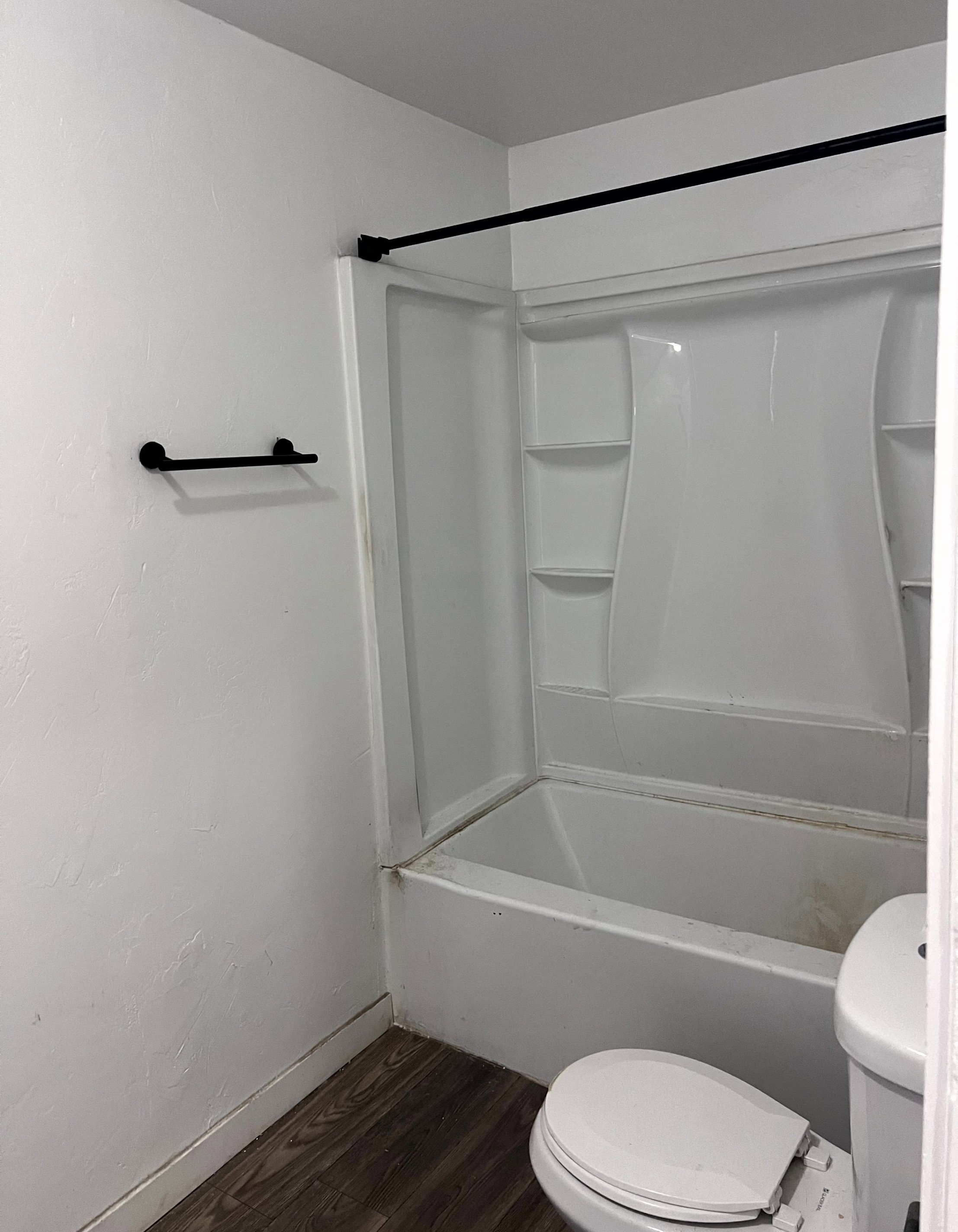 Bathroom featuring a white tub-shower combination unit, matte black shower rod, matte black towel bar, and wood-finish flooring