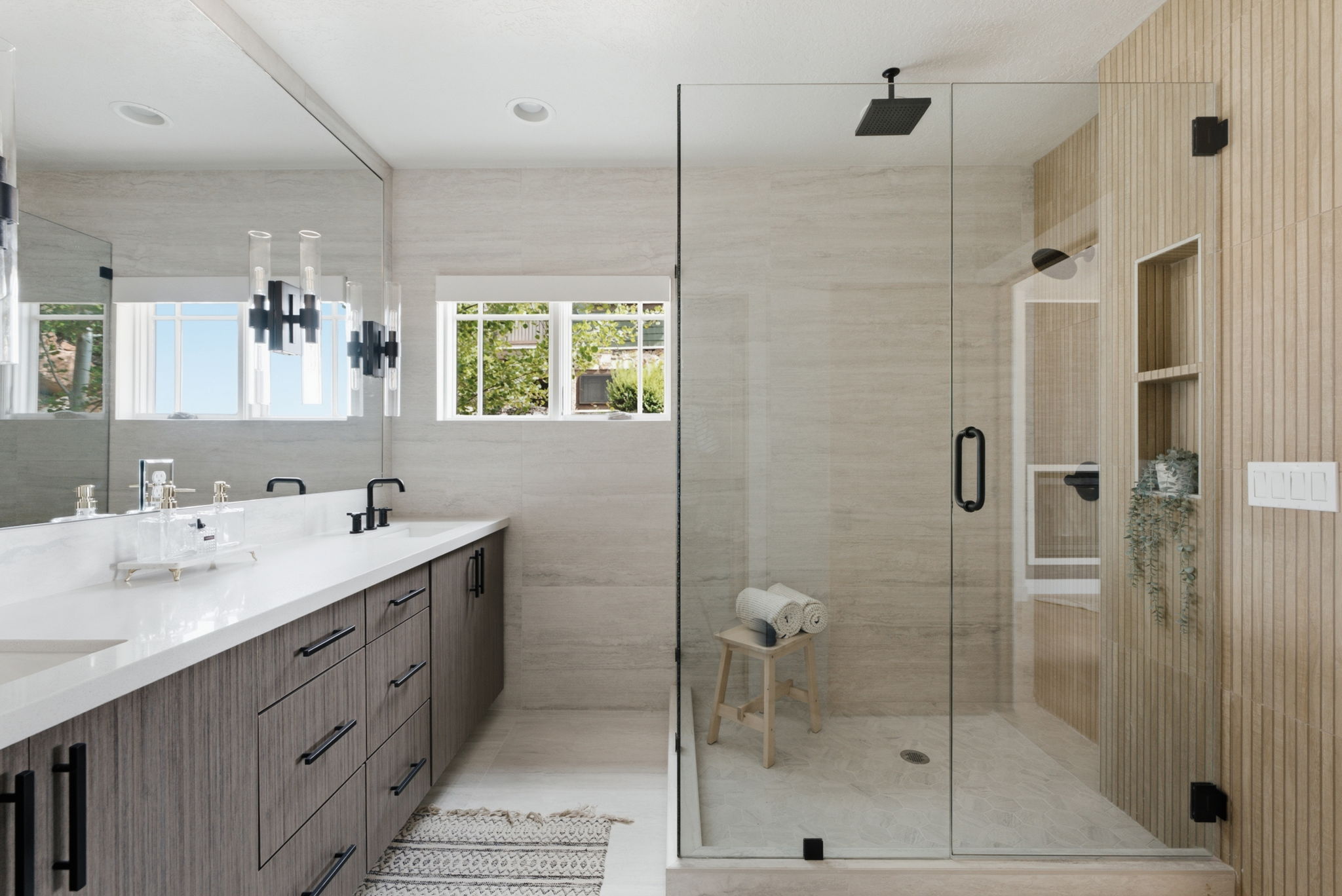 Modern bathroom featuring a glass-enclosed shower with a matte black rain showerhead, a dual-sink vanity with a white countertop and wood-finish cabinetry, and light-toned wall and floor tile
