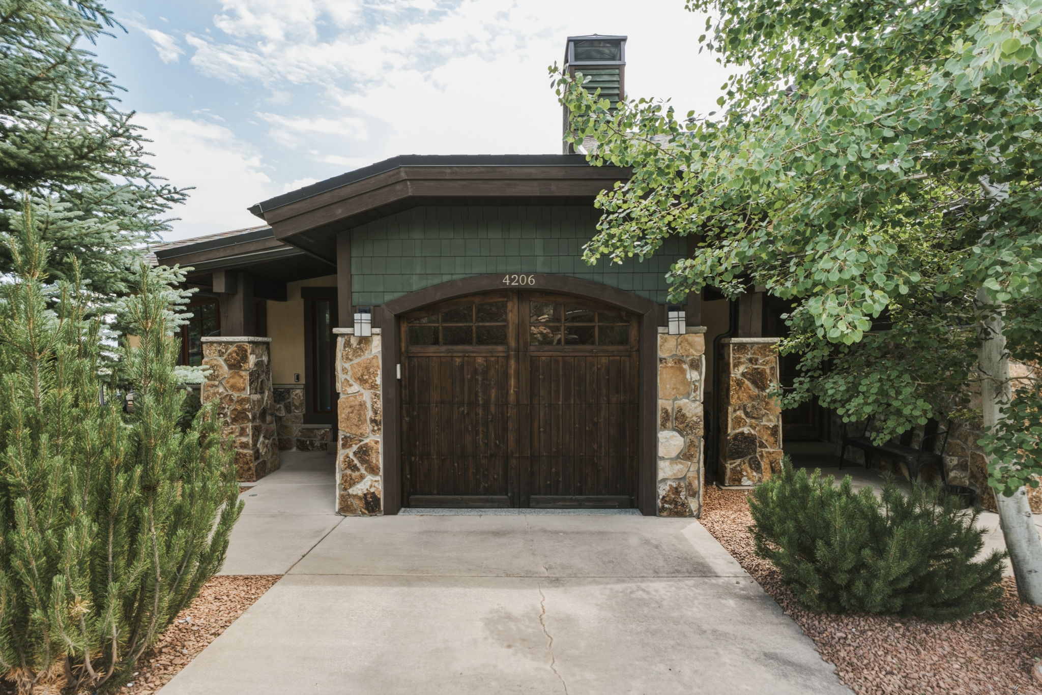Craftsman-style exterior featuring stone columns, a wood-paneled garage door with an arched top, green shingle siding, and a prominent chimney