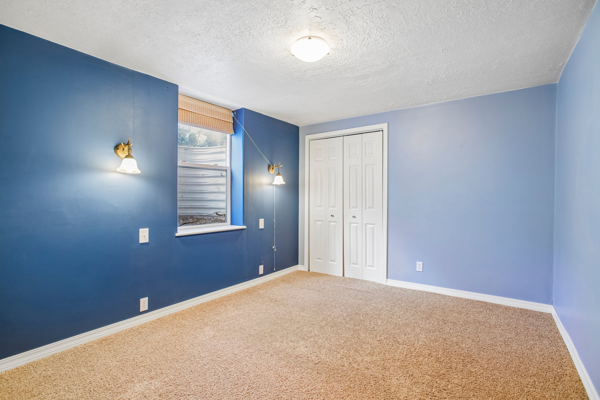 Carpeted room featuring a window with a natural wood-finish blind, dual bifold closet doors, and two wall-mounted sconce fixtures