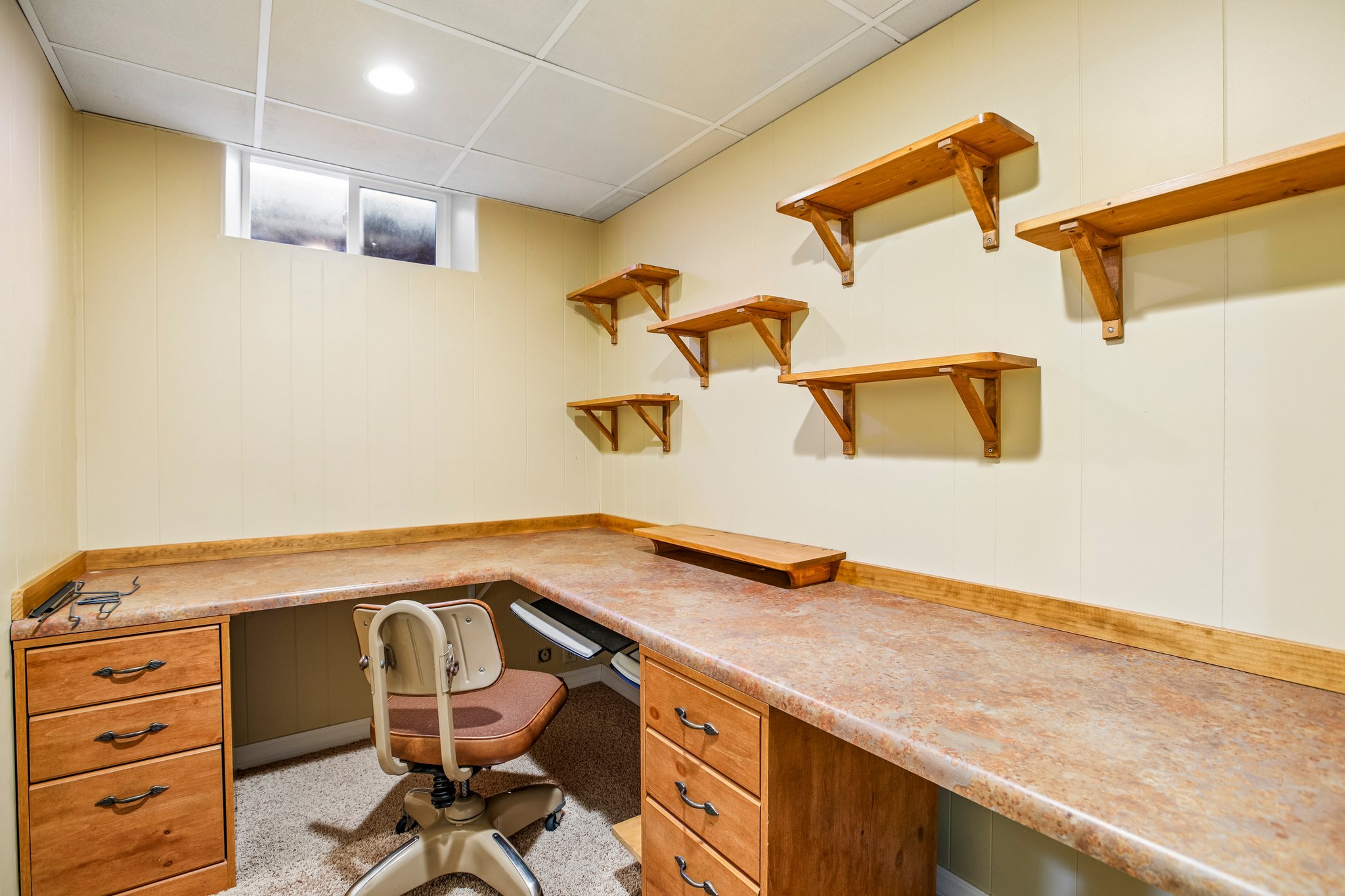 Room featuring extensive built-in L-shaped desk with multiple drawers, light-colored paneling, recessed lighting, and a series of wall-mounted wood shelves