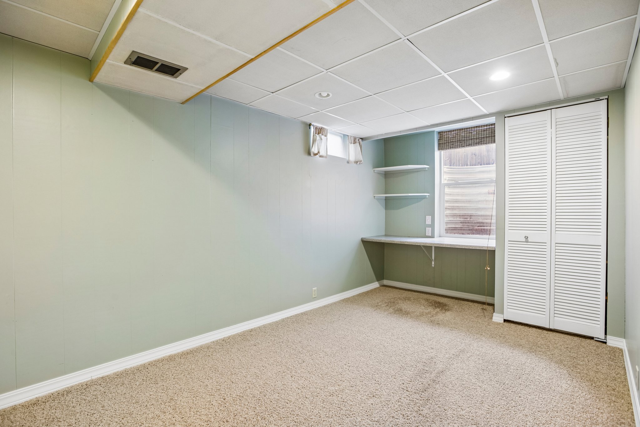Carpeted room featuring a drop ceiling with recessed lighting, paneled walls, a built-in desk with shelving, and louvered bi-fold closet doors