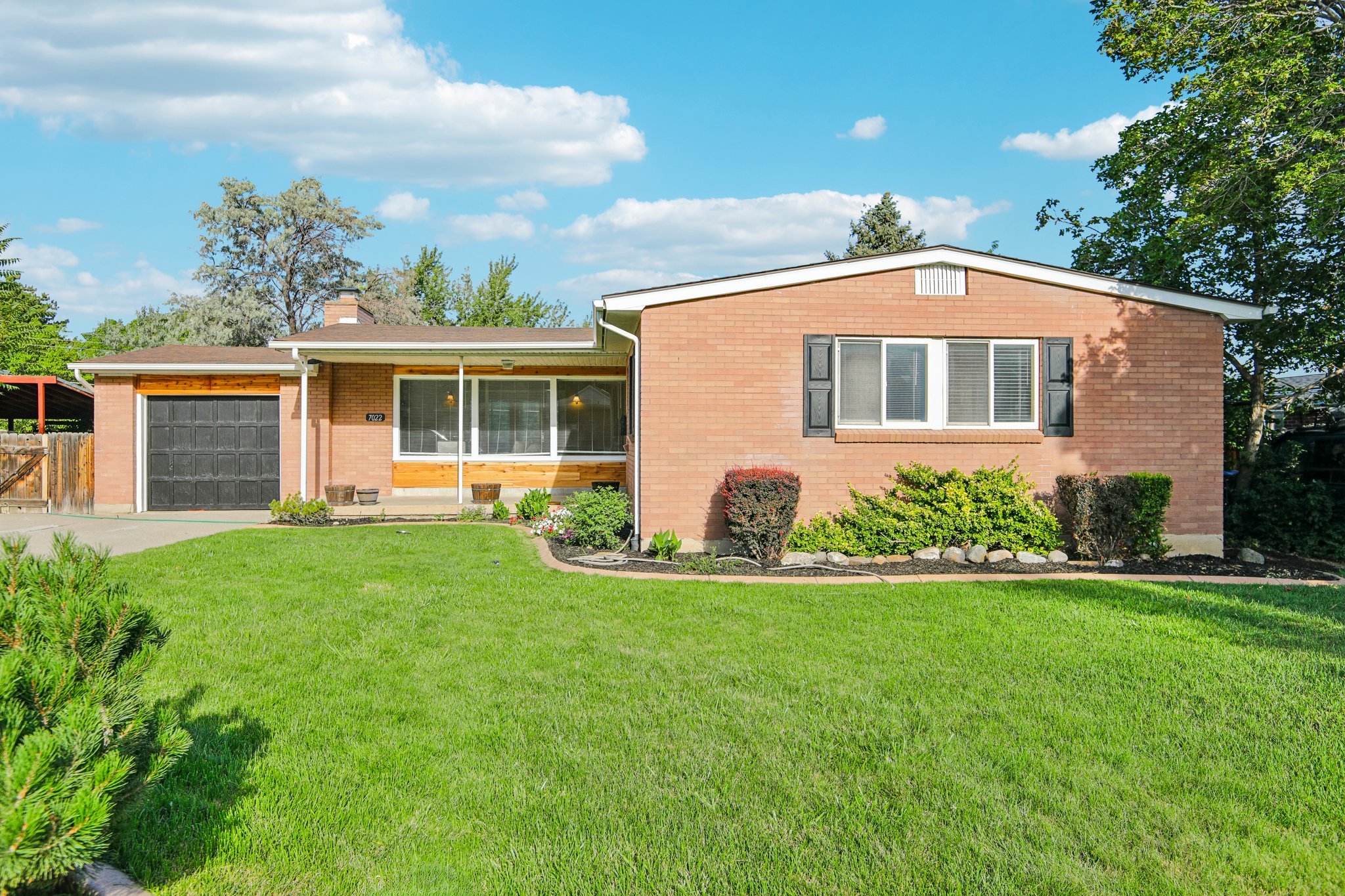Brick ranch home featuring a front-facing garage with a dark door, a manicured lawn, and mature landscaping
