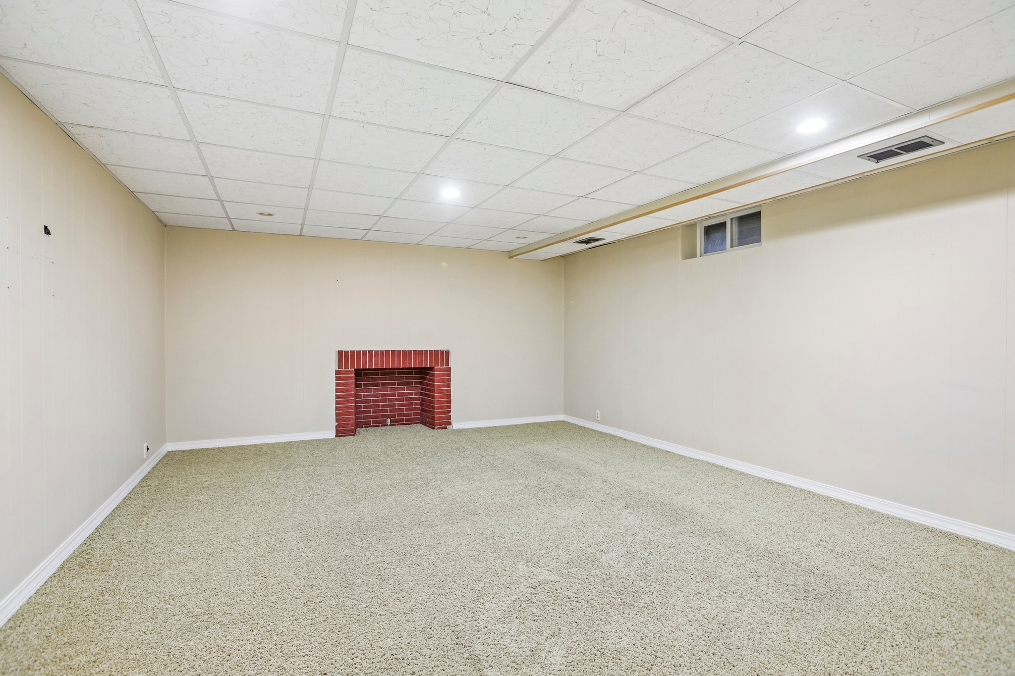 Spacious room featuring a red brick fireplace, light-toned carpeting, white baseboards, and a grid panel ceiling with recessed lighting