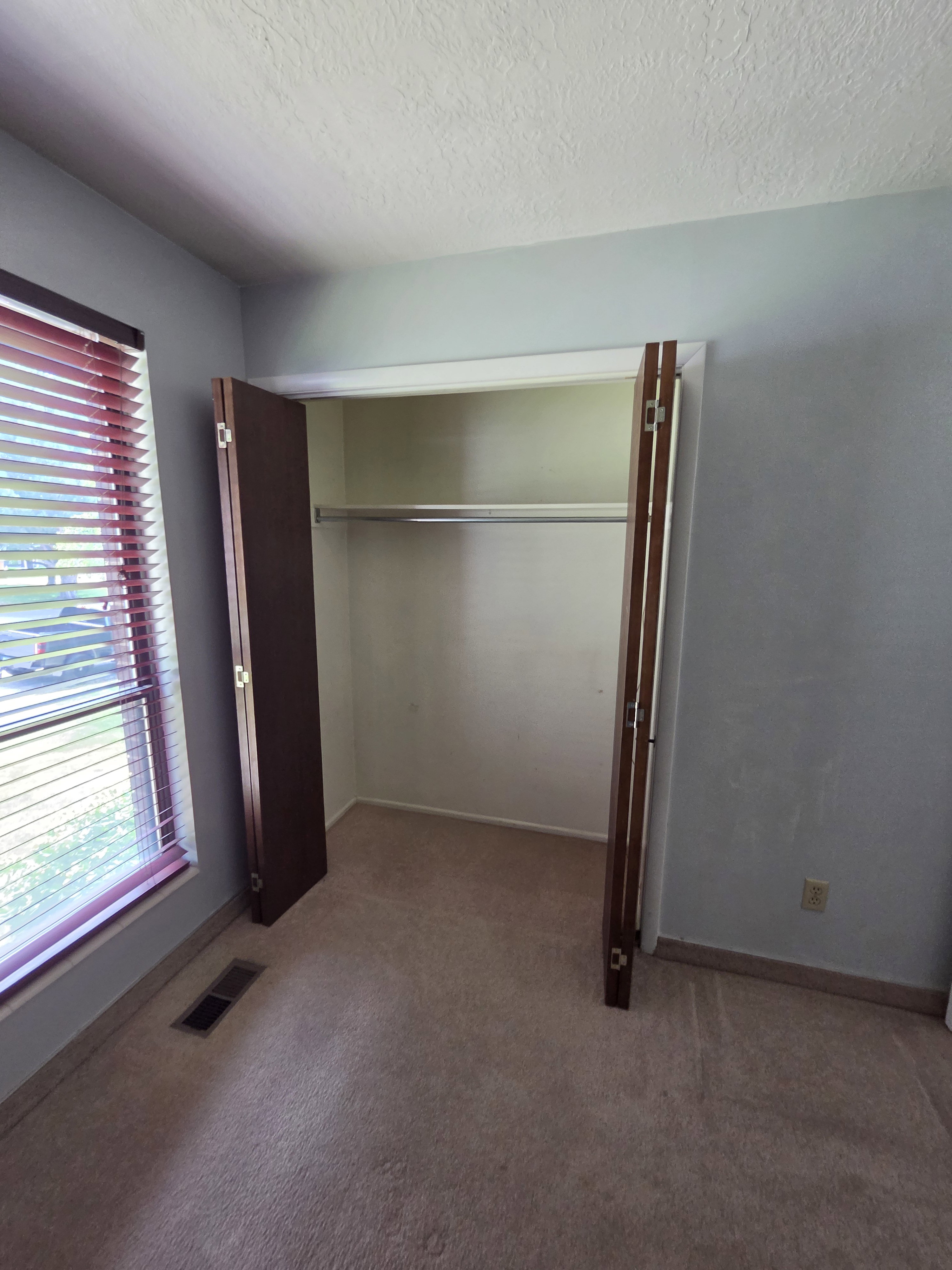 Recessed closet with bi-fold doors and a built-in hanging rod, carpeted flooring, and a window with horizontal blinds