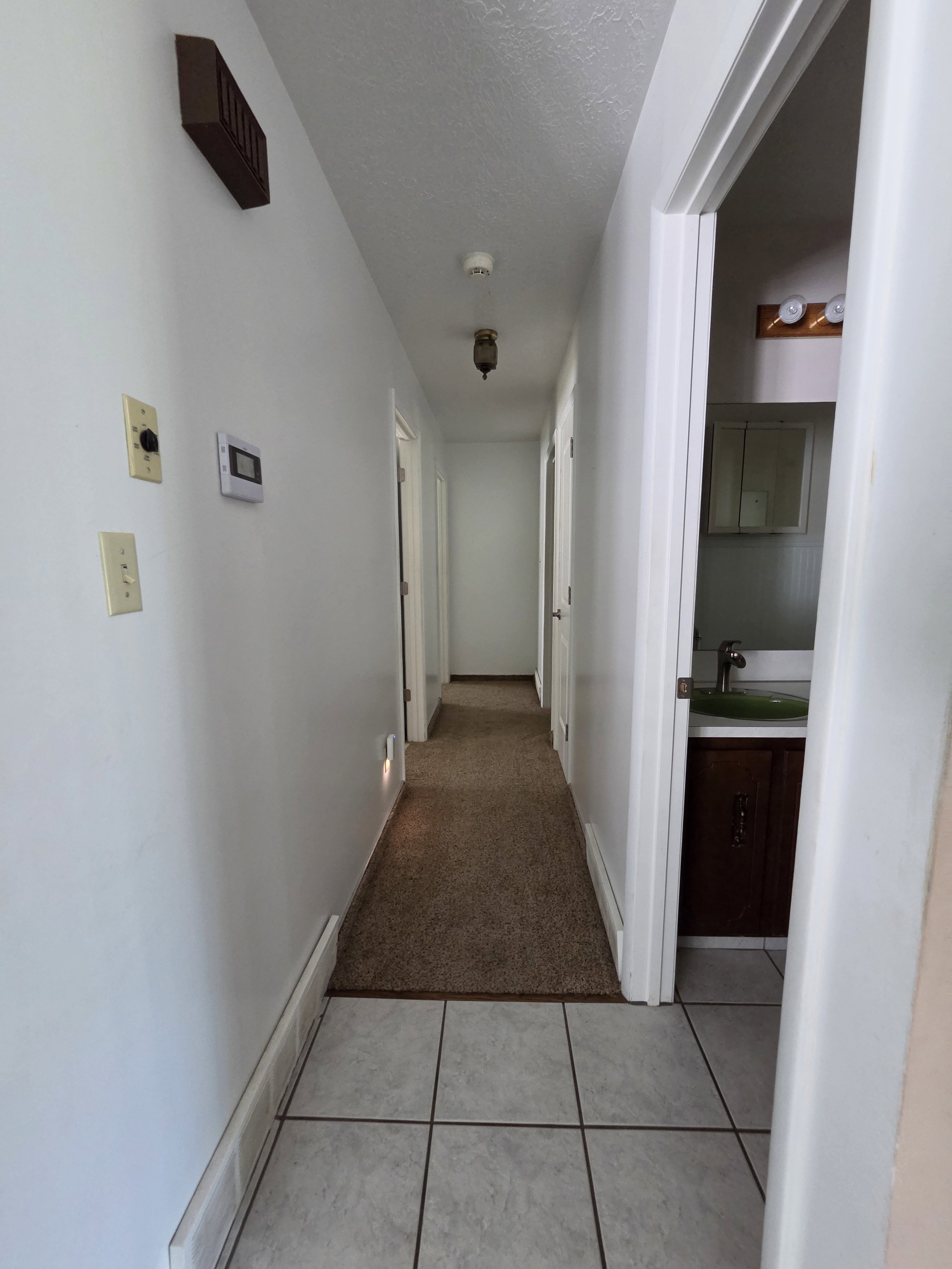 Hallway featuring tile flooring transitioning to carpet, white walls, and a ceiling-mounted light fixture