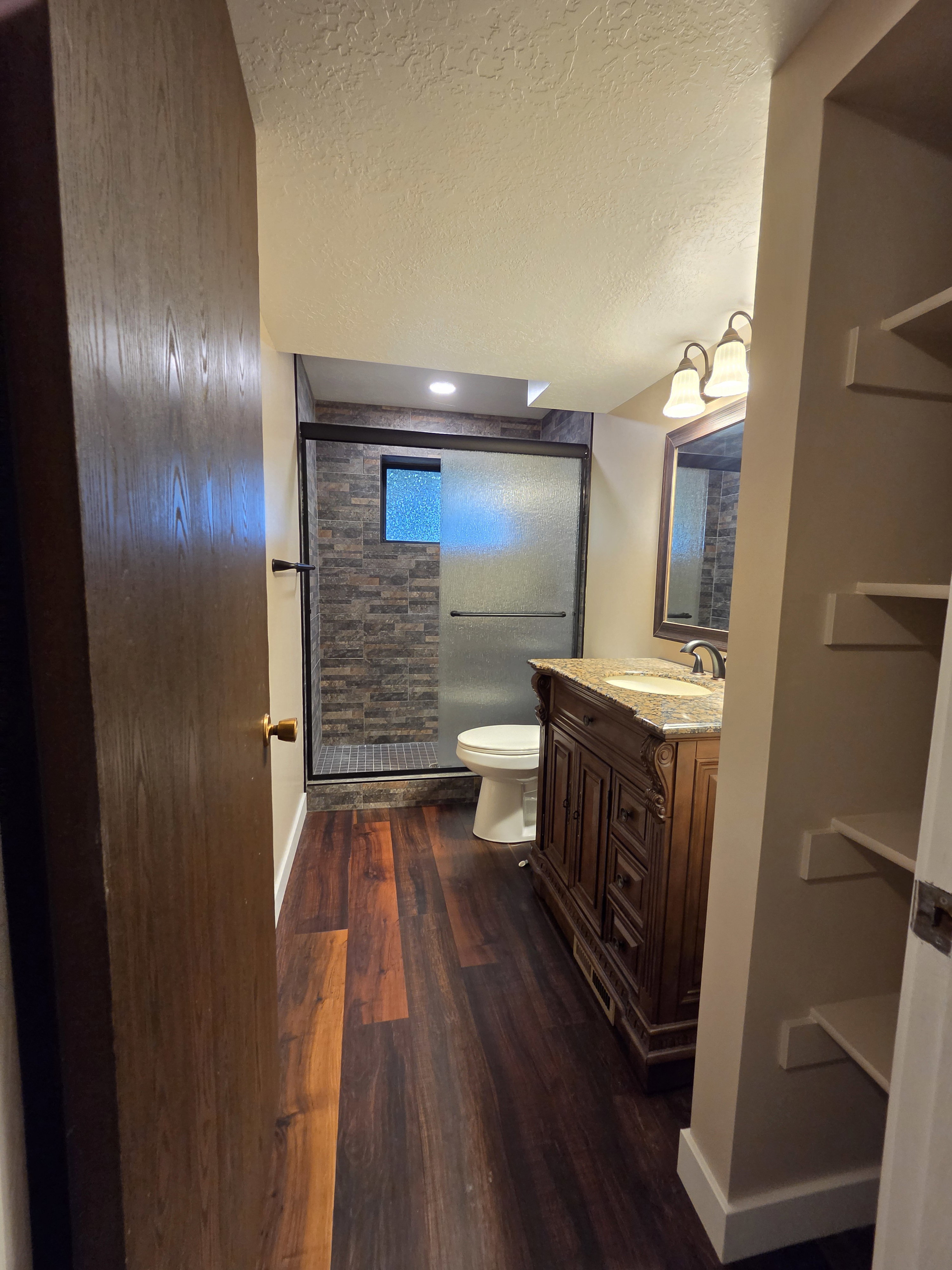 Spacious bathroom featuring dark wood-finish flooring, a decorative vanity with a built-in sink, a framed mirror, and a walk-in shower with stone-look wall tile and a frosted glass enclosure