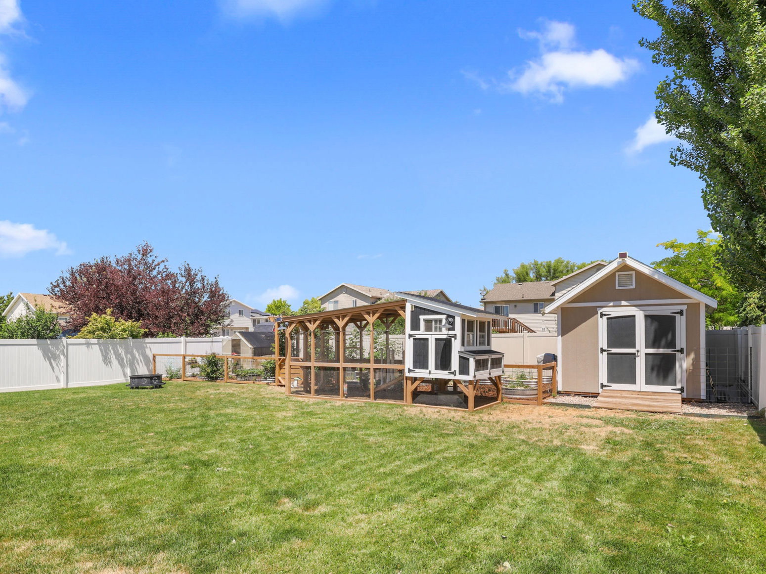 Expansive green lawn featuring a substantial storage shed with double doors, an enclosed chicken coop structure, and a perimeter white vinyl fence