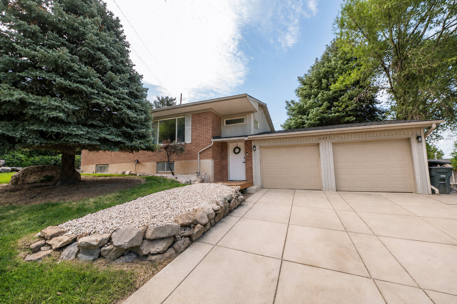 Brick facade home featuring a two-car garage, concrete driveway, stone retaining wall, and mature trees