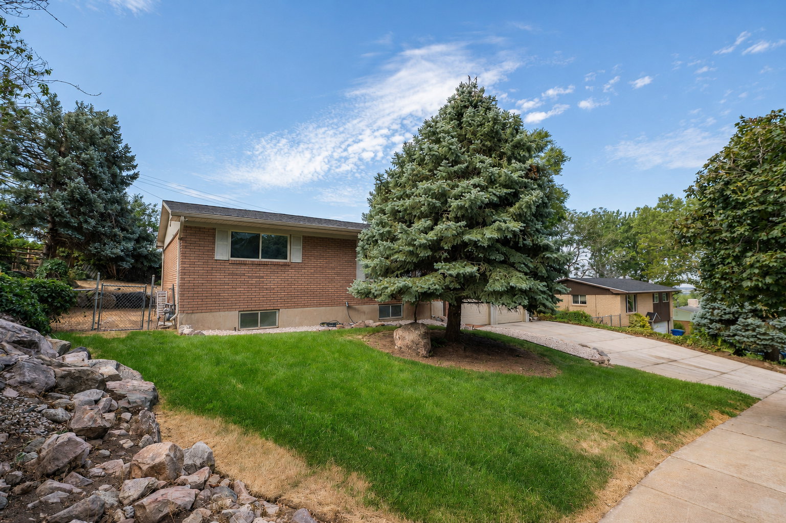 Brick exterior home featuring a well-maintained front lawn, mature evergreen tree, paved driveway, and established landscaping