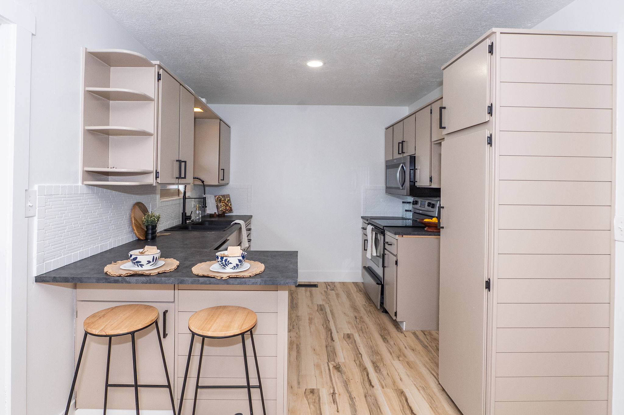 Contemporary kitchen featuring wood-finish flooring, light-toned cabinetry with black hardware, dark countertops, a white subway tile backsplash, and stainless steel appliances including a microwave and range