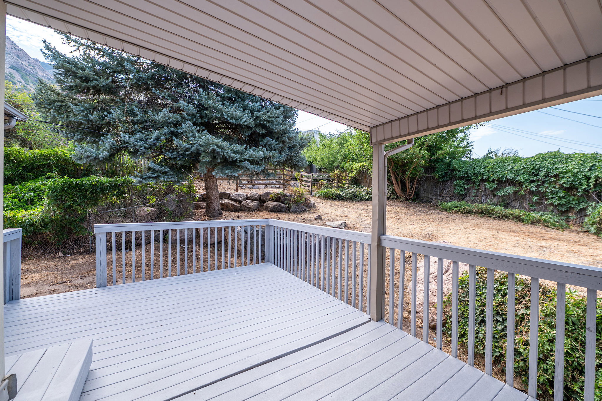 Covered patio deck with light gray wood-finish decking and railing overlooking a sloped backyard with mature trees and natural rock landscaping