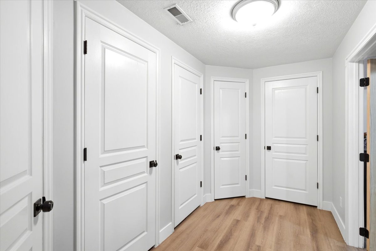 Interior hallway featuring storage closets with five-panel doors with matte black hardware, light wood-finish flooring, white baseboards, and a flush-mount ceiling light fixture