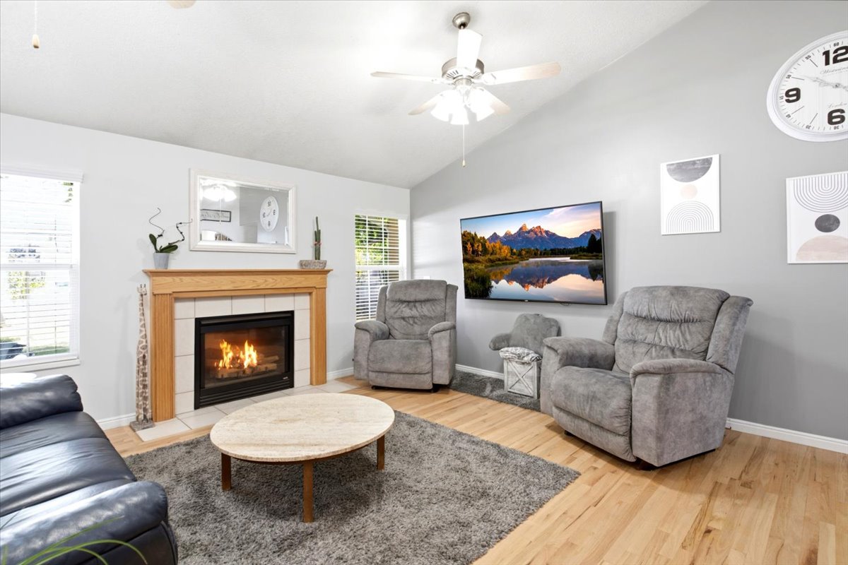 Living area featuring a fireplace with a wood-finish mantle and tile surround, vaulted ceiling, two windows with blinds, and wood-finish flooring