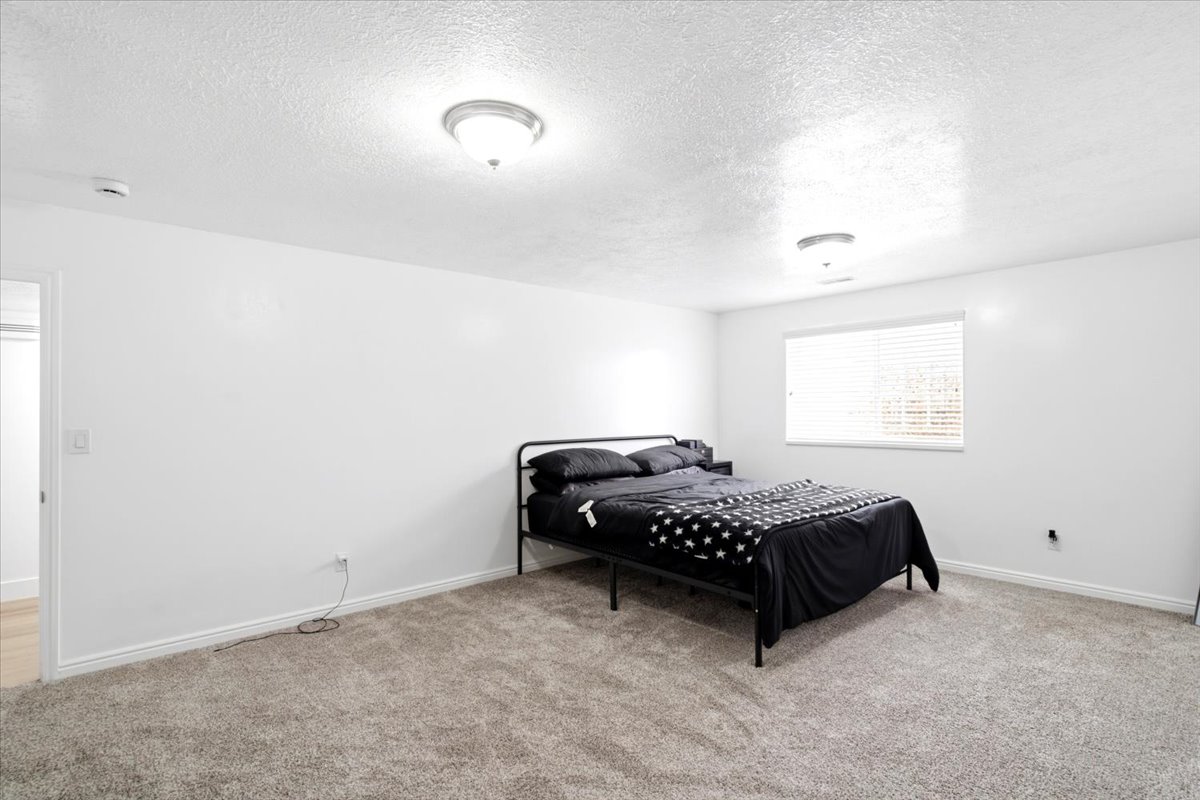 Spacious room with light gray carpeting, crisp white walls, two ceiling-mounted light fixtures, and a window with horizontal blinds