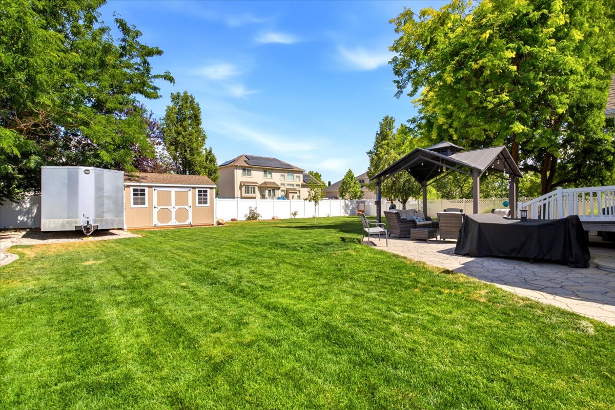 Expansive backyard featuring a lush lawn, dedicated patio with a gazebo, white privacy fencing, a storage shed, and mature trees