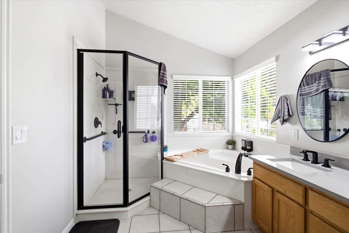 Spacious Primary bathroom featuring a glass-enclosed shower with black trim, a built-in bathtub with tiled surround, dual windows, a single vanity with wood-finish cabinetry, and a round mirrors