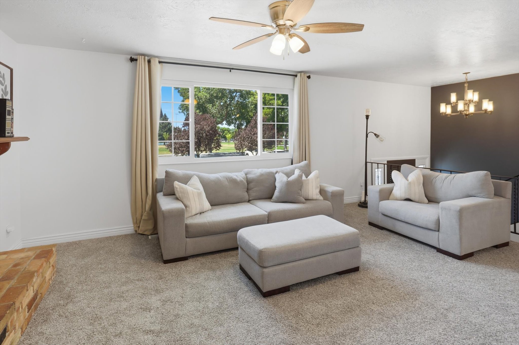 Open living space featuring a large window, light-colored carpeting, a ceiling fan, a brick hearth, and a chandelier