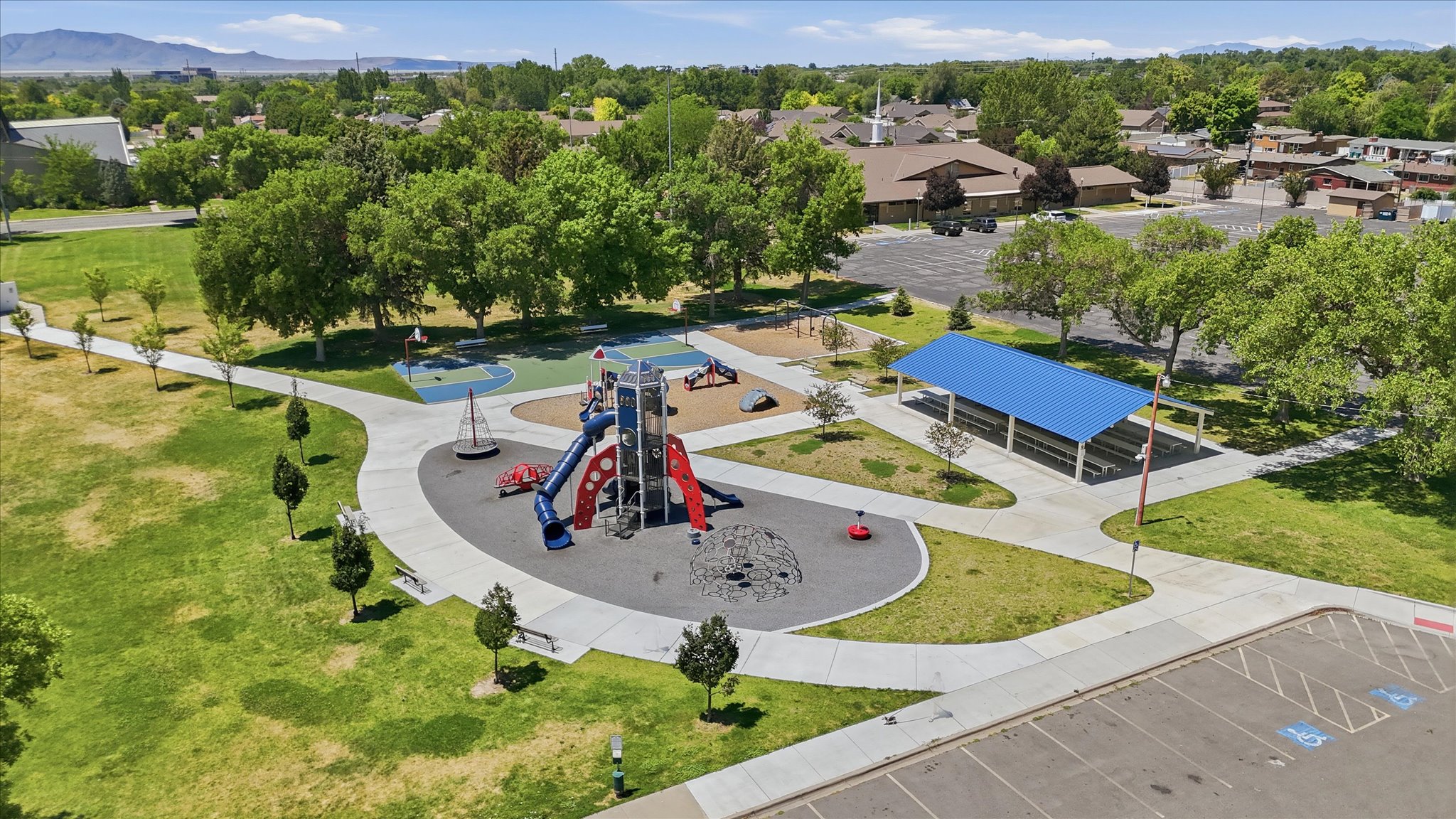 Outdoor recreation area featuring a modern playground structure with slides, a covered picnic area, a basketball court, and expansive green spaces