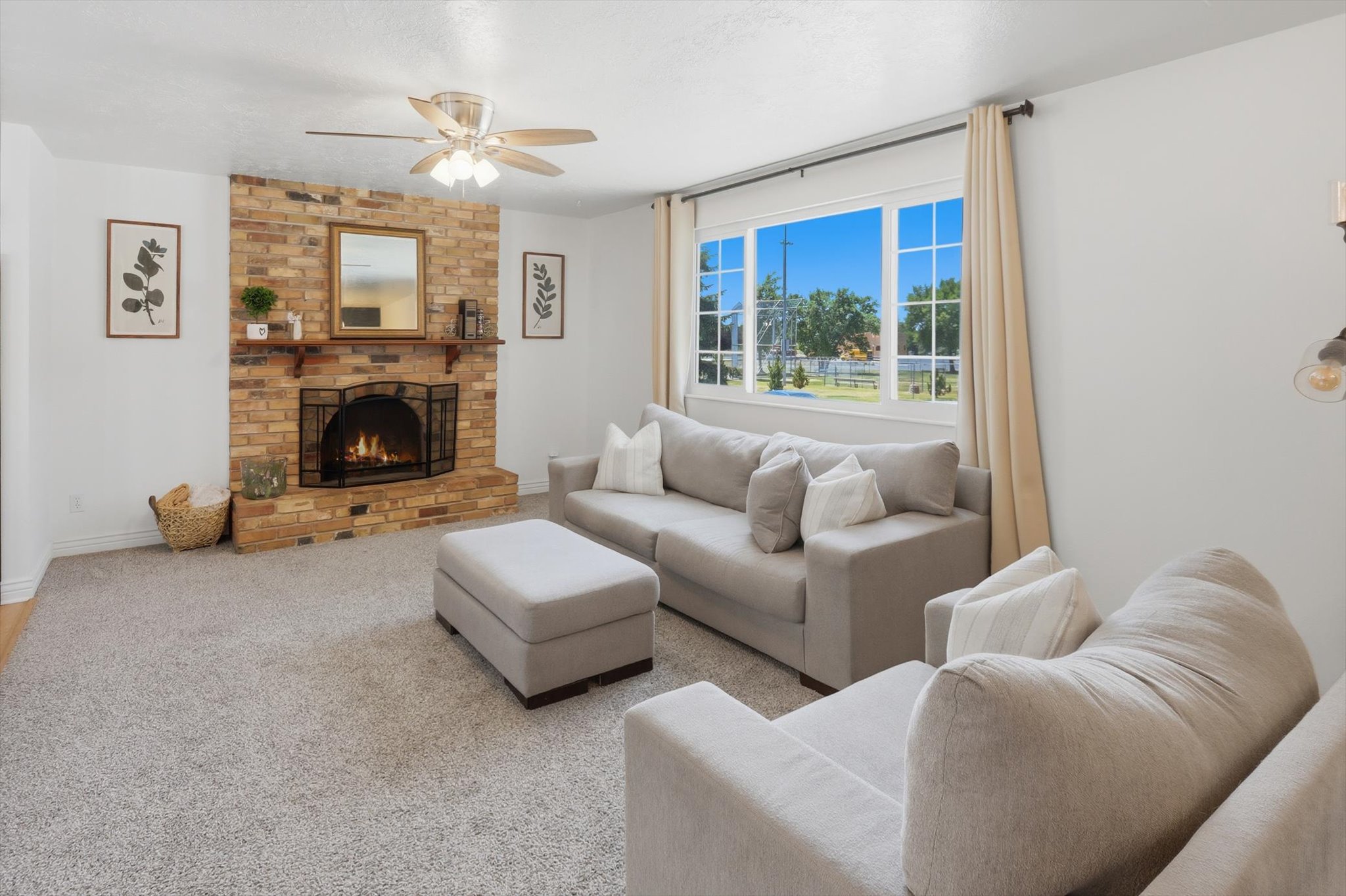 Living area featuring a brick fireplace with a wood mantle, a ceiling fan, a large multi-pane window with drapes, and light-toned carpeting