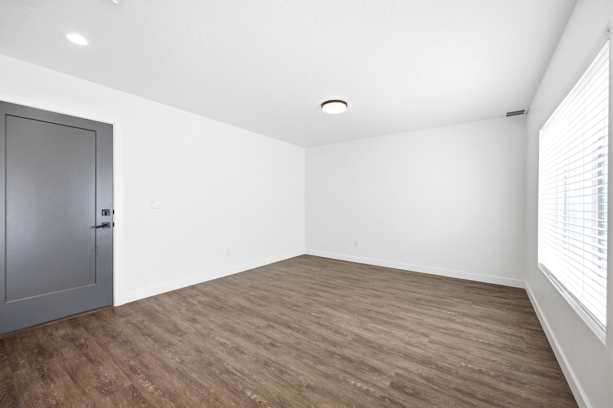 Spacious room featuring wood-finish flooring, white walls, and a large window with blinds