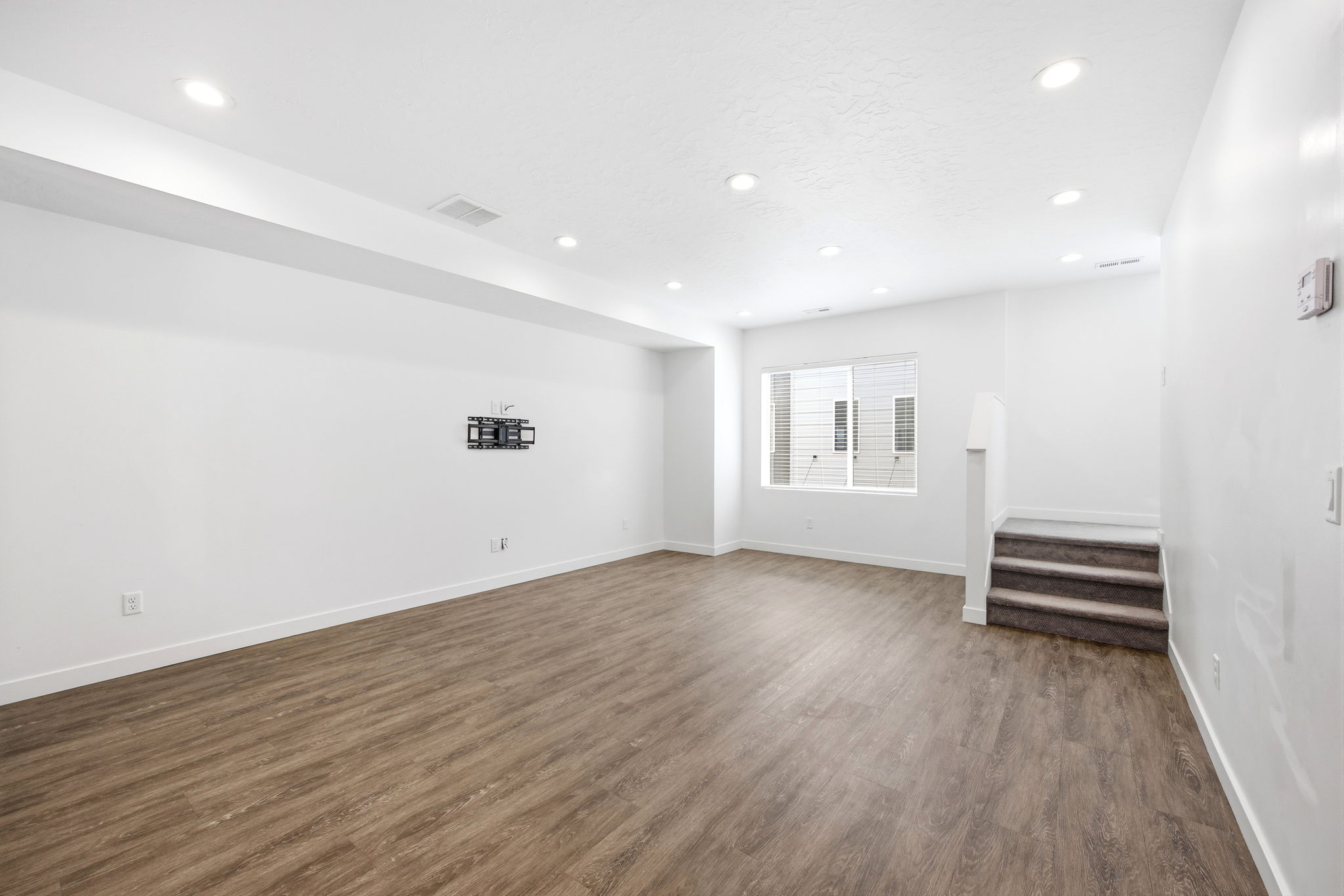 Spacious lower-level room featuring wood-finish flooring, recessed lighting, white baseboards, and a window providing natural illumination