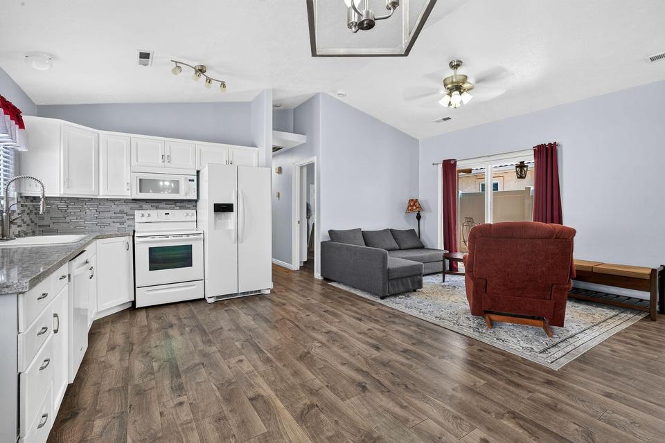 Open-concept interior featuring wood-finish flooring throughout, vaulted ceilings, and a kitchen with white cabinetry, granite-style countertops, and a tile backsplash
