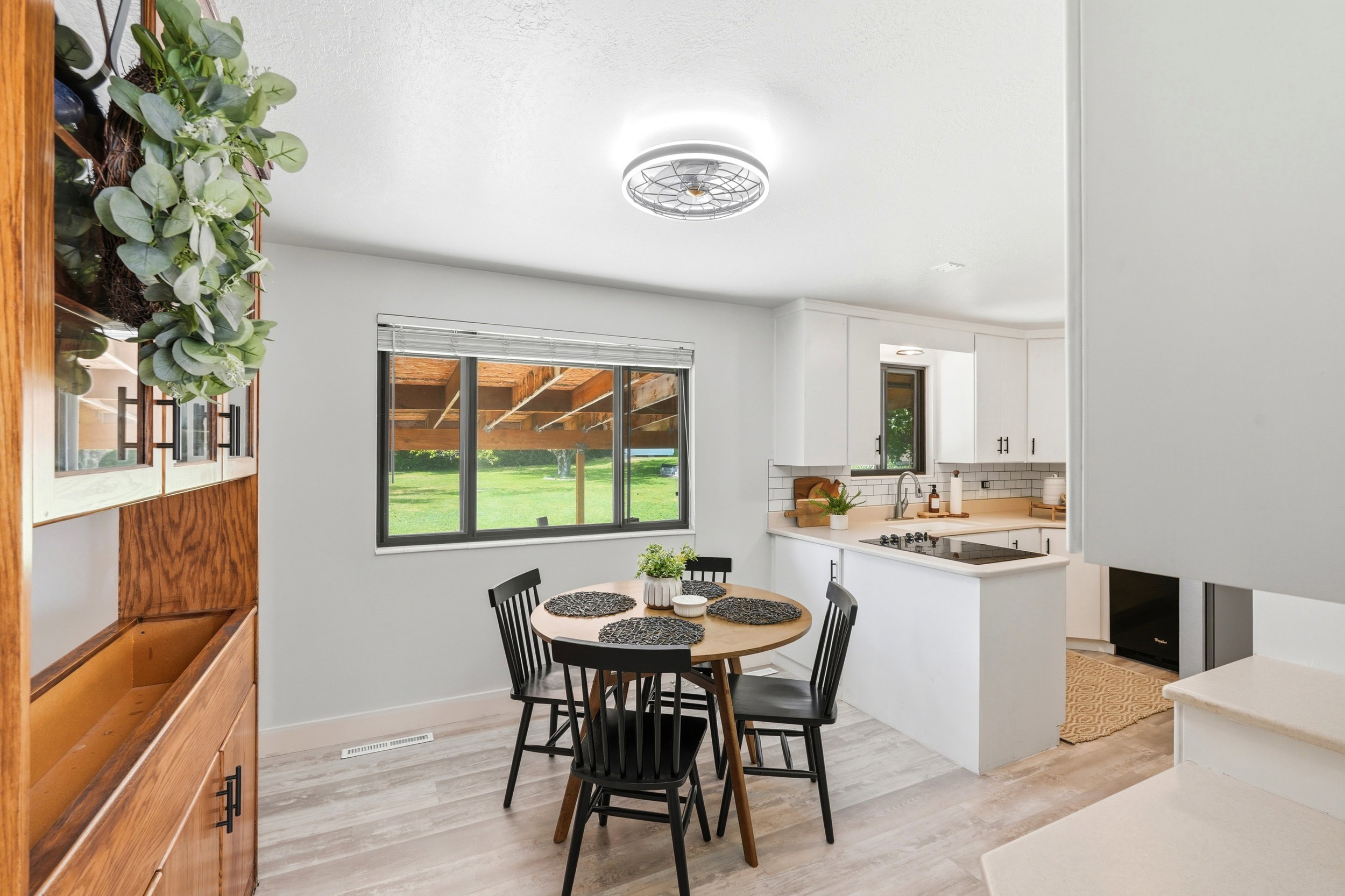 Open concept living area featuring a kitchen with white cabinetry and a light-toned countertop, a dining area with wood-finish flooring, and a large window overlooking a green outdoor space