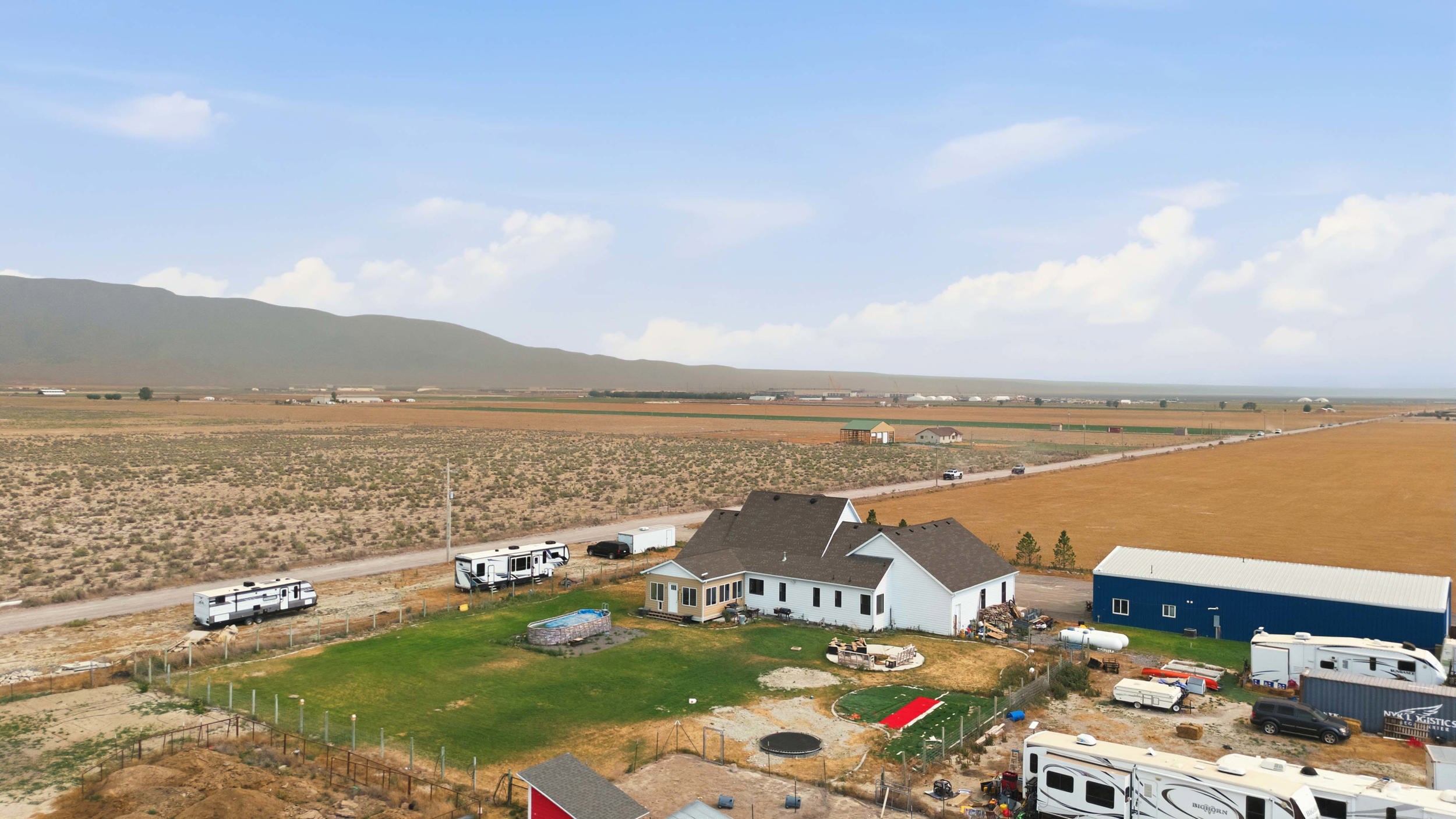 Expansive property featuring a white residence with a brown shingled roof, a large blue outbuilding, a green lawn, and substantial undeveloped land extending to distant mountains