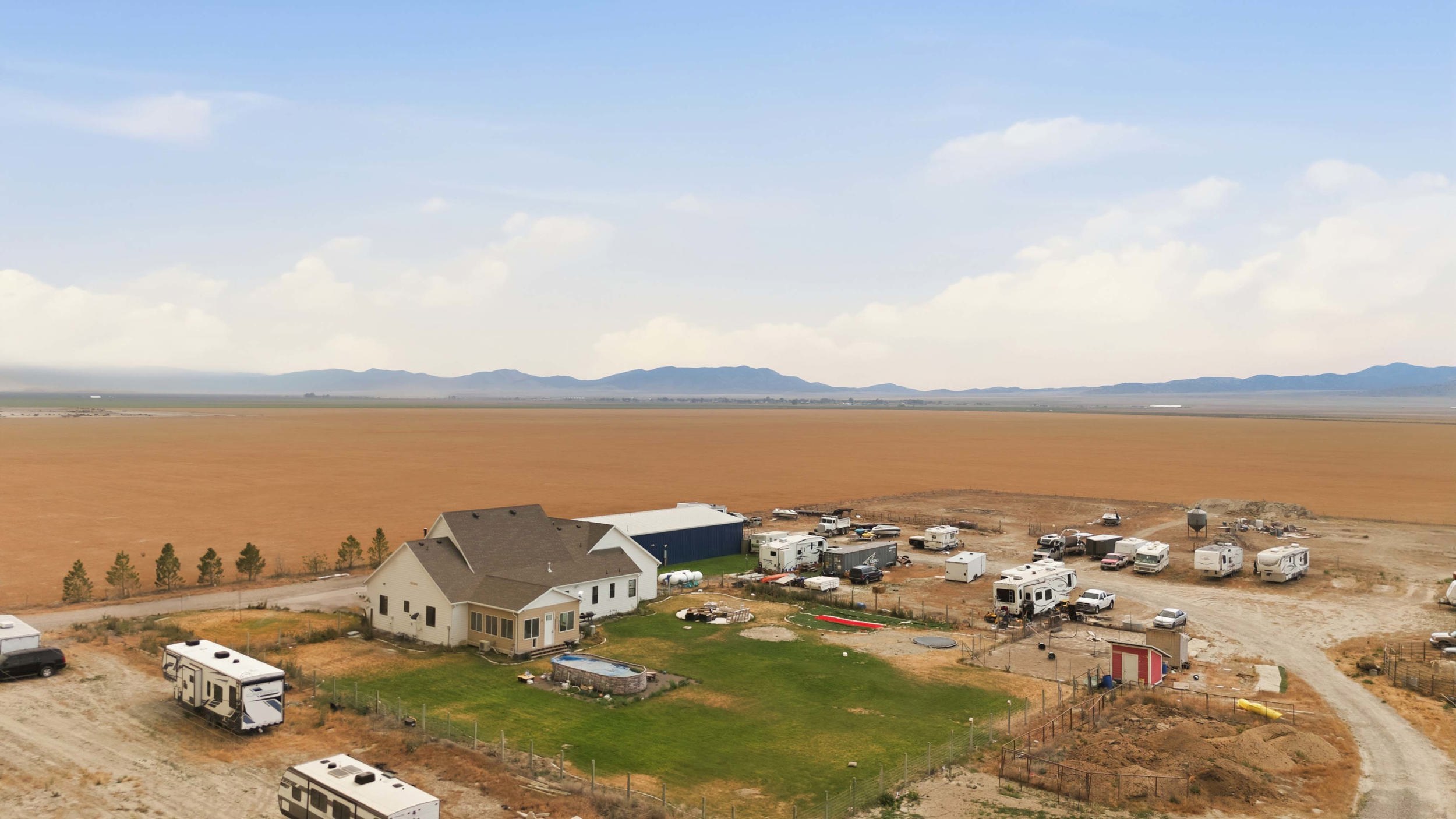 Expansive rural property featuring a beige stucco residence with a brown shingle roof, a blue-sided outbuilding, and a large open field backdrop