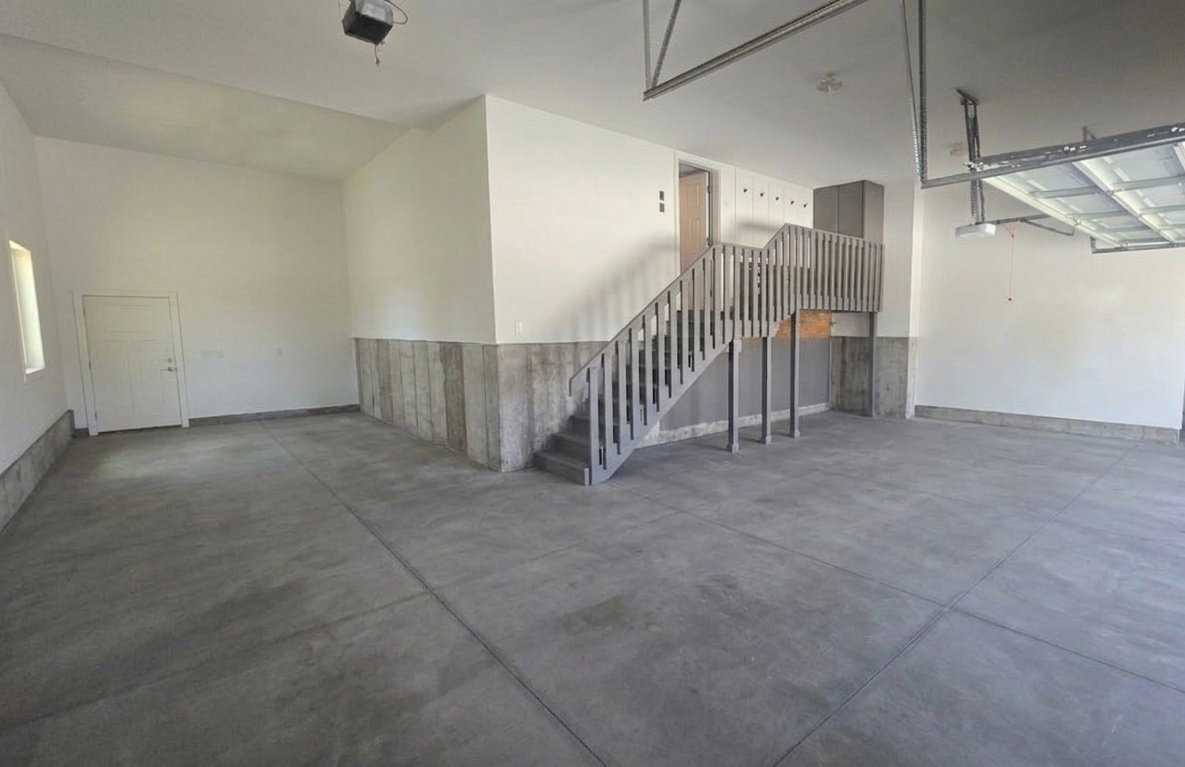 Spacious garage featuring a concrete floor, painted white walls, and a finished staircase with wooden railings