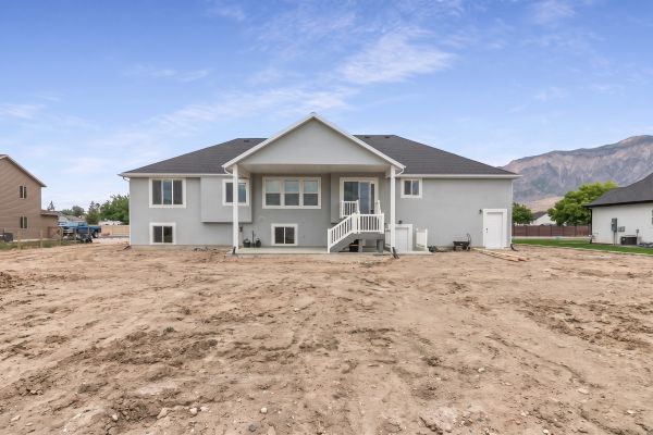 Spacious rear exterior featuring a light gray stucco finish, black shingle roofing, multiple windows, and a sliding glass door leading to a covered patio
