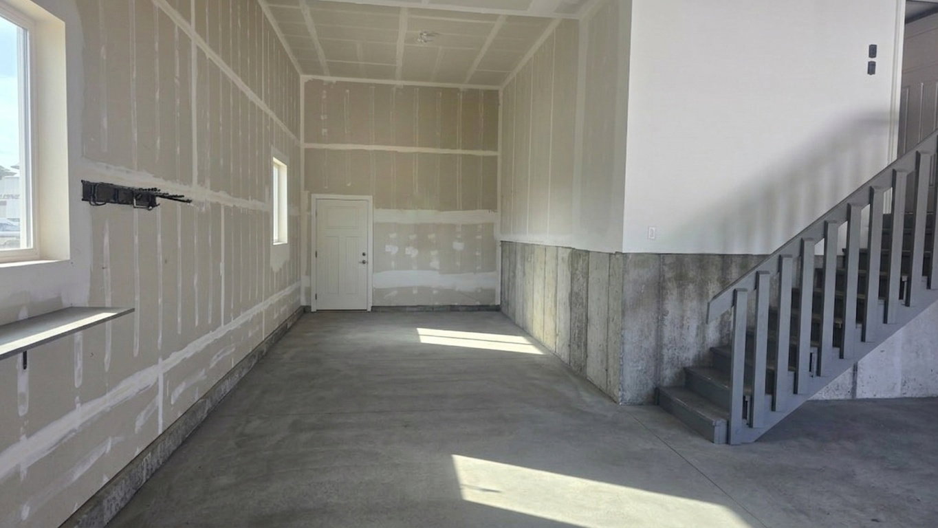 Spacious unfinished garage with an exposed concrete floor, interior access door, large window, and staircase featuring a painted railing