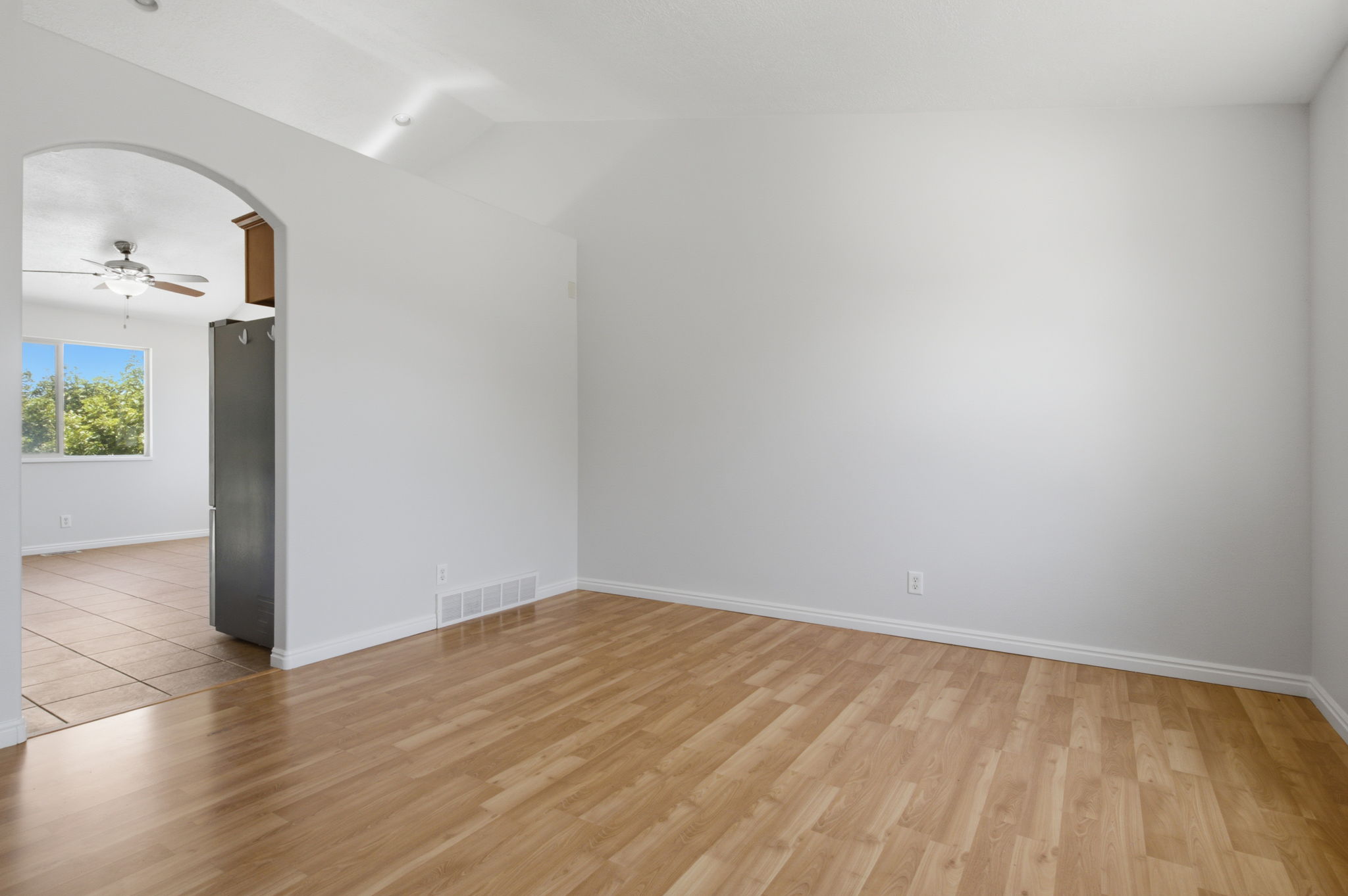 Vaulted ceilings and wood-finish flooring characterize this open space, featuring an arched entryway to an adjoining room with tile flooring and a ceiling fan