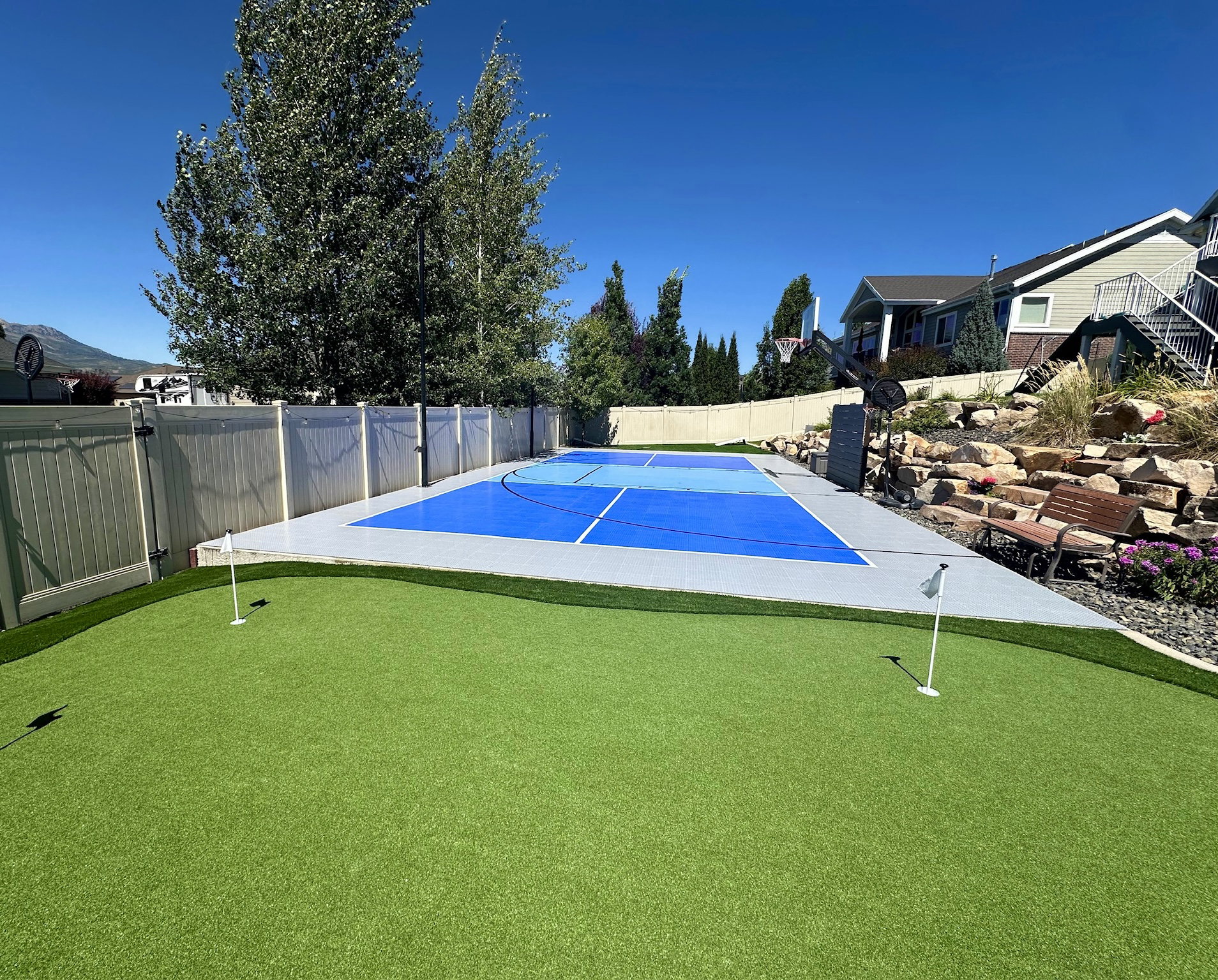 Backyard featuring a synthetic turf putting green with two flags, a multi-sport court with blue surfacing, and a rockery with integrated landscaping