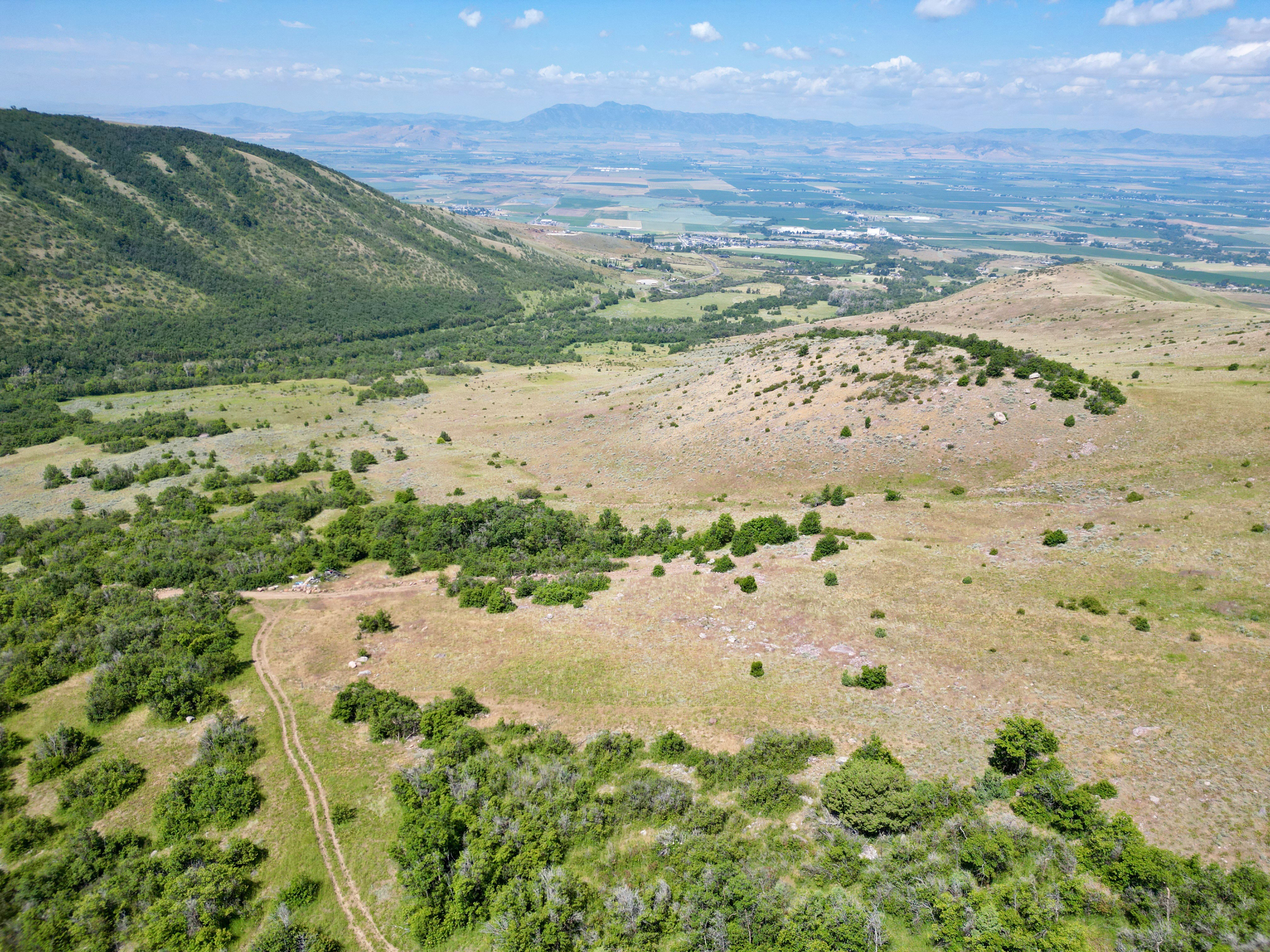 Expansive mountainous terrain with varied vegetation, featuring a prominent valley and distant agricultural lands under a bright sky