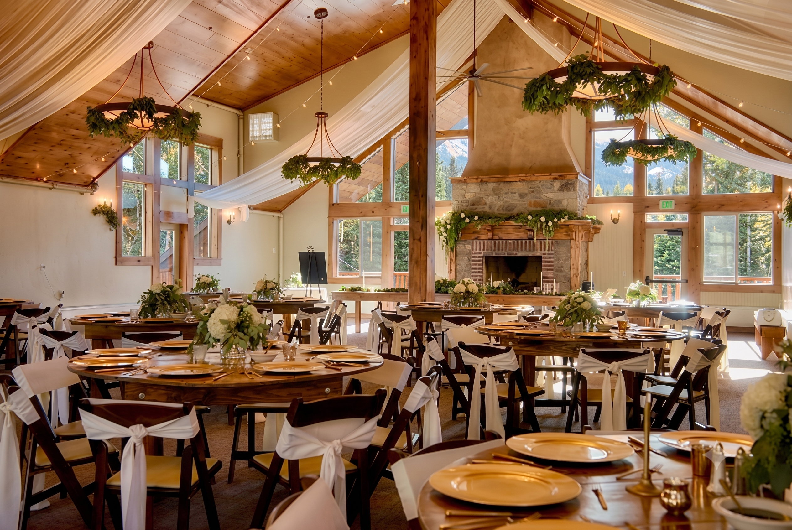 Spacious interior featuring a prominent stone fireplace, vaulted wood-paneled ceilings, large windows, exposed structural beams, and decorative chandeliers