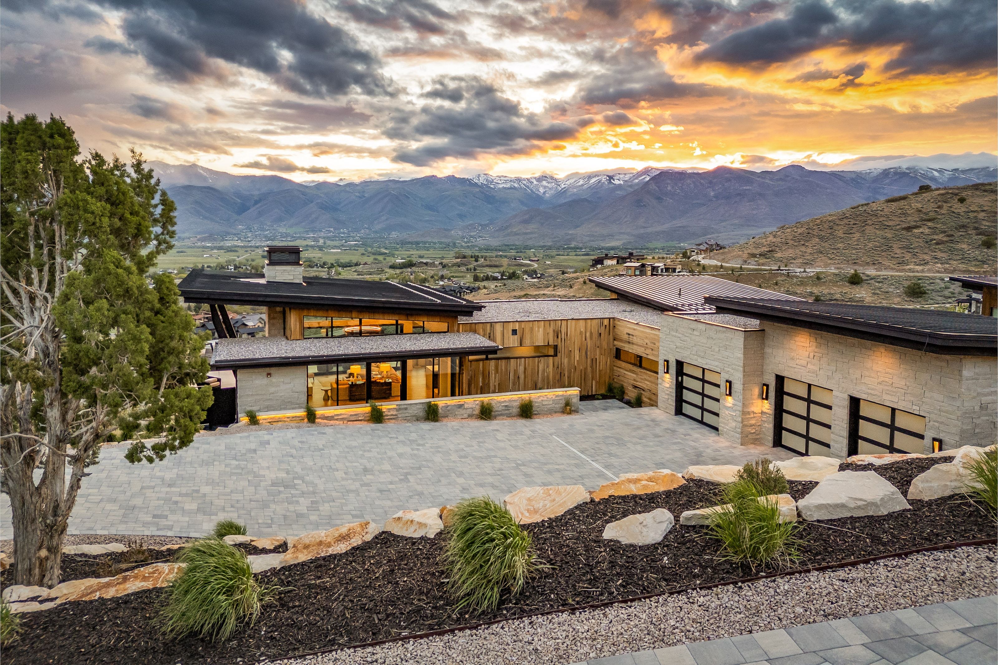 Contemporary residence featuring a stone and wood-finish exterior, expansive glass windows, a paved driveway, and three modern garage doors