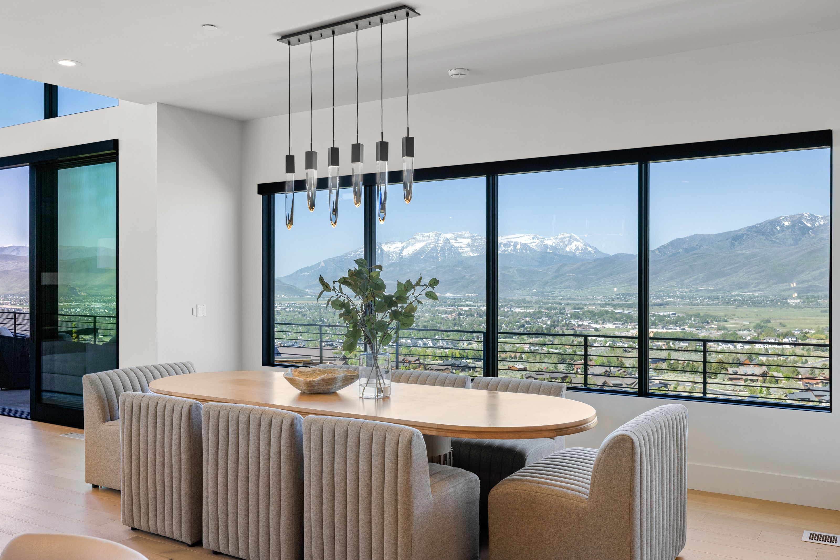 Expansive window wall offering a mountain and city scenery, complemented by light wood-finish flooring, a modern linear chandelier, and a large sliding glass door to exterior
